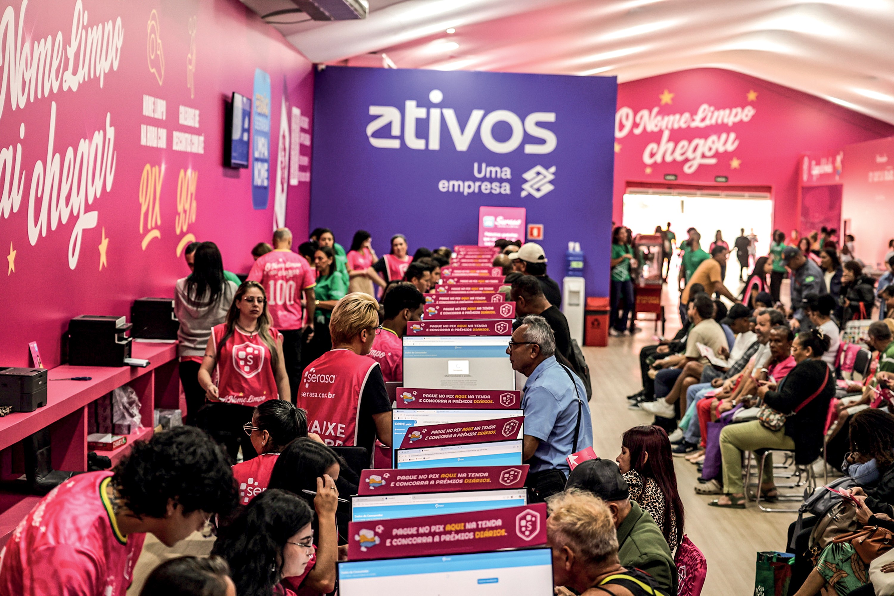 Pessoas em um evento de serviços financeiros, com paredes rosa e roxas. Muitos participantes estão sentados em frente a computadores, enquanto outros circulam. A parede roxa exibe o logo ativos Uma empresa e a rosa, O Nome Limpo chegou.
