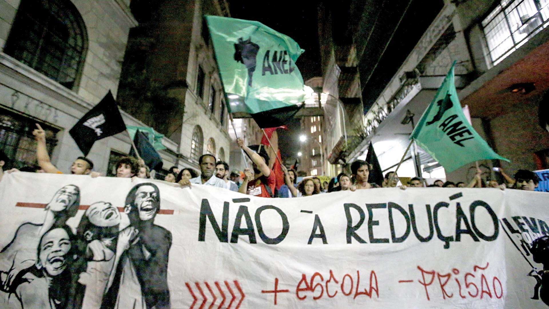 Manifestantes seguram uma faixa branca com a frase NÃO À REDUÇÃO em letras pretas e + ESCOLA - PRISÃO em vermelho. Pessoas diversas, algumas com bandeiras verdes da ANEL e pretas, participam do protesto em uma rua urbana com edifícios ao fundo