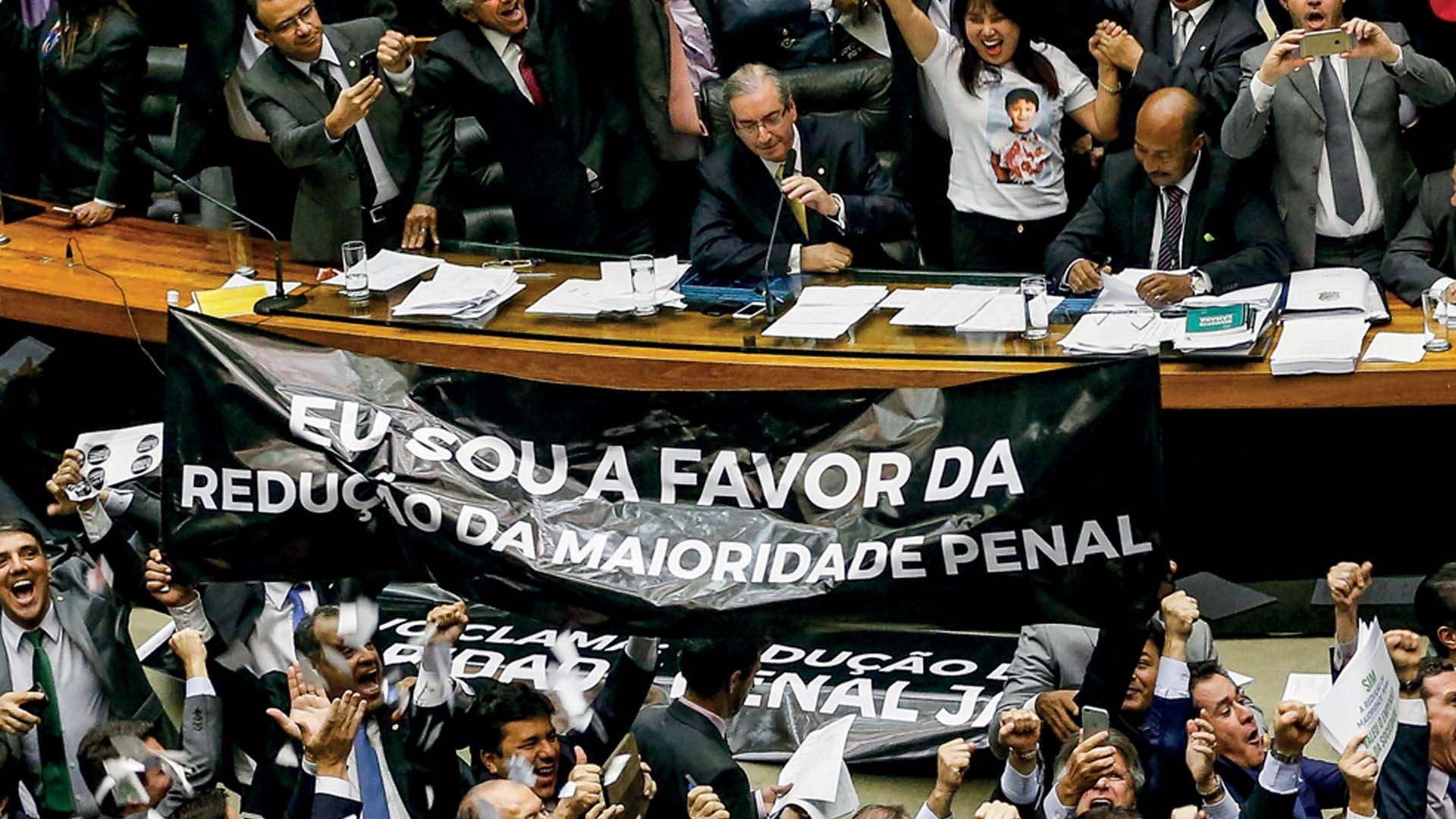 Políticos em plenário, alguns de terno, outros com camisas de protesto, exibem uma faixa preta com a frase EU SOU A FAVOR DA REDUÇÃO DA MAIORIDADE PENAL em letras brancas. Alguns sorriem, outros gesticulam, e um homem de óculos e terno cinza está sentado à mesa, olhando para baixo.