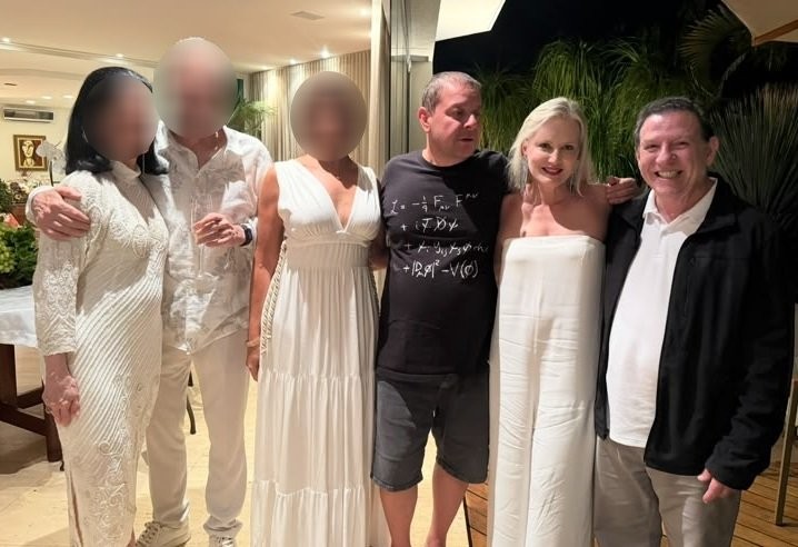 Seis pessoas, três mulheres e três homens, posam para foto em ambiente interno. À esquerda, uma mulher de vestido branco longo e um homem de camisa branca abraçam uma mulher de vestido branco. Ao centro, um homem de camiseta preta com fórmulas e bermuda escura abraça uma mulher loira de macacão branco tomara que caia. À direita, um homem sorridente de camisa polo branca e jaqueta preta. O fundo mostra uma sala com iluminação indireta e plantas
