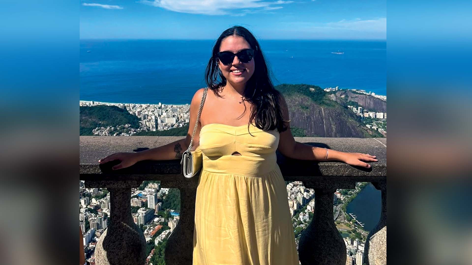 Mulher sorridente de cabelos escuros e óculos de sol, vestindo um vestido tomara que caia amarelo, apoiada em um parapeito de pedra. Ao fundo, a cidade do Rio de Janeiro com prédios, montanhas e o mar azul sob céu claro