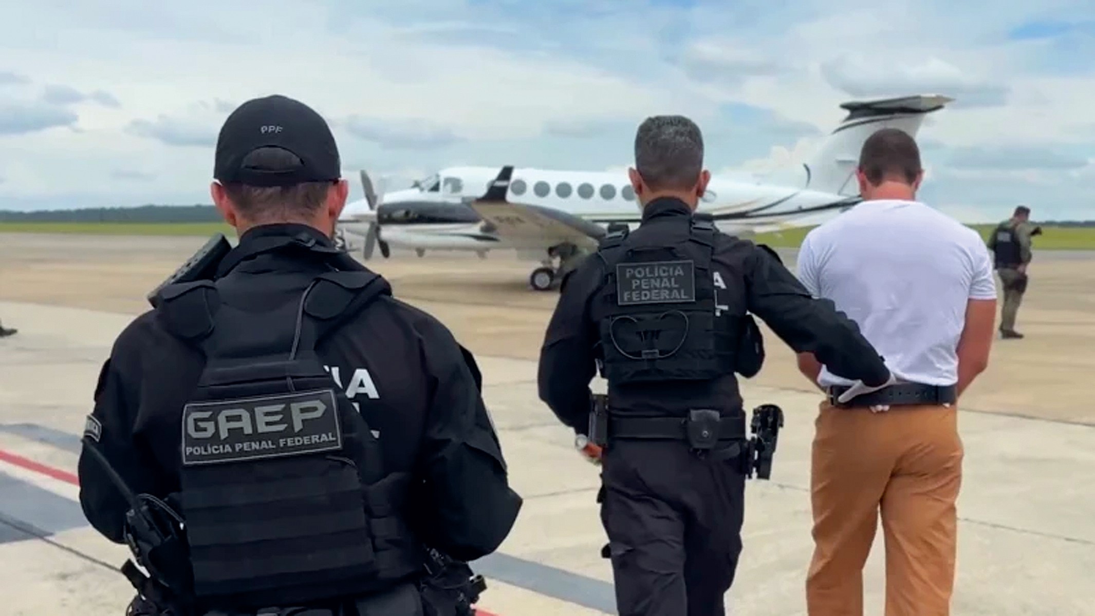 Três homens, dois policiais penais federais e um detento algemado, caminham em uma pista de aeroporto com um avião branco ao fundo