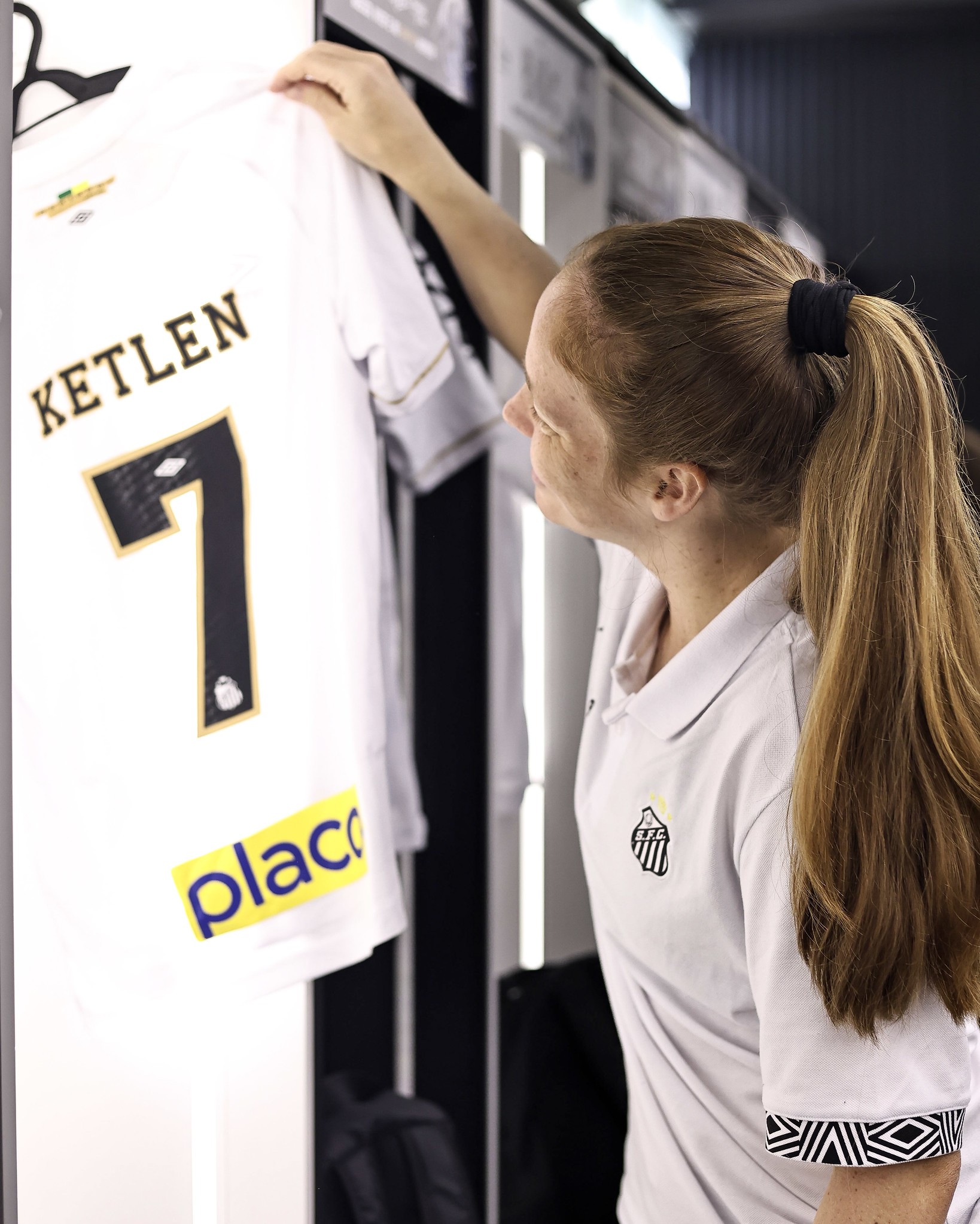Mulher de cabelos ruivos presos em rabo de cavalo, vestindo camisa polo branca do Santos, segura uma camisa de futebol branca com o nome KETLEN e o número 7 em preto e dourado, em um vestiário
