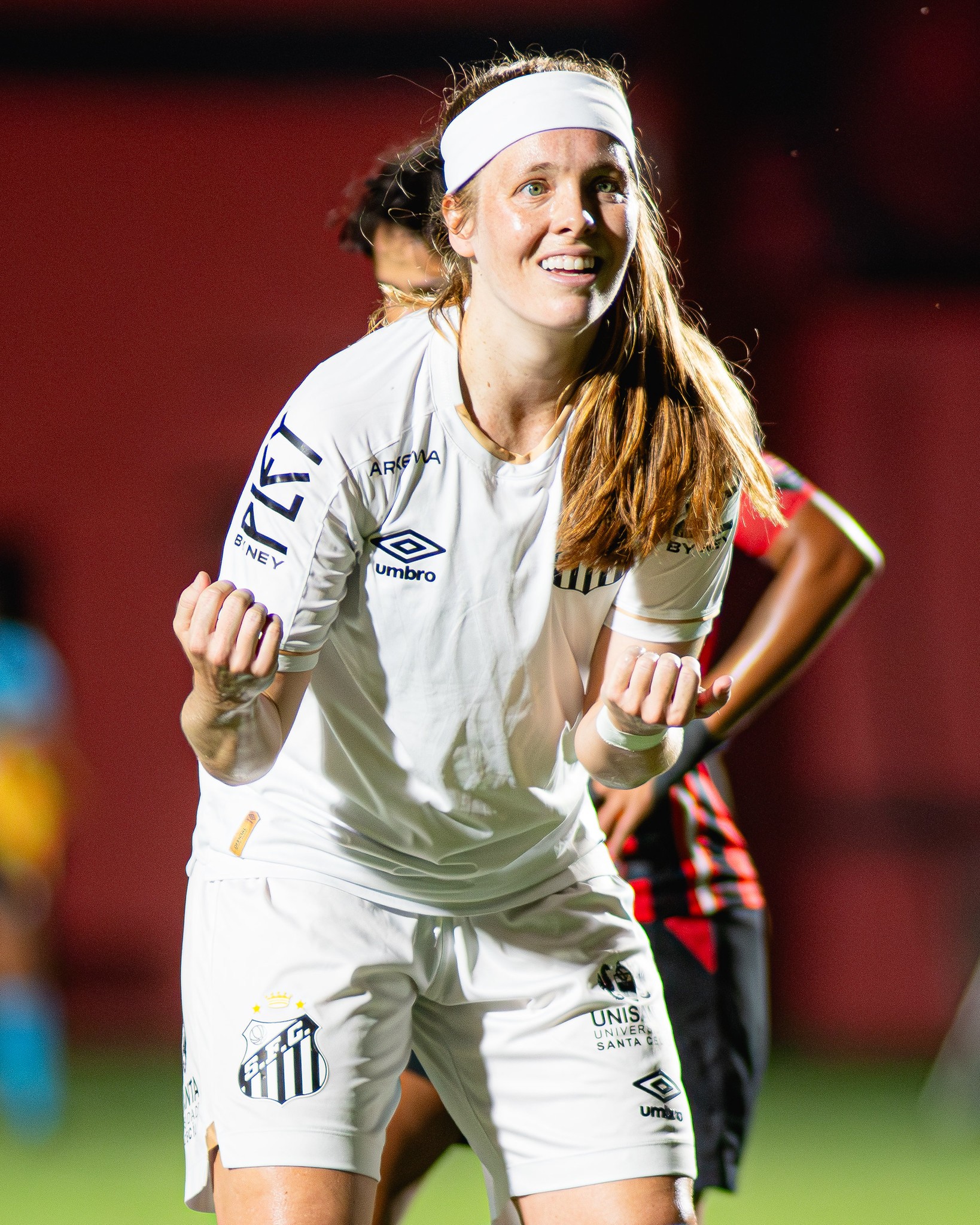 Jogadora de futebol feminina do Santos, loira de bandana branca, sorri e celebra com os punhos cerrados, vestindo uniforme branco com detalhes pretos, em campo de futebol à noite