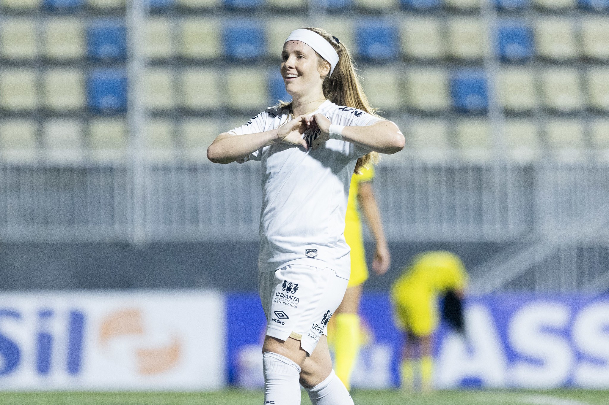 Jogadora de futebol feminina, loira, sorrindo e fazendo um coração com as mãos, vestindo uniforme branco em campo de futebol com arquibancada ao fundo