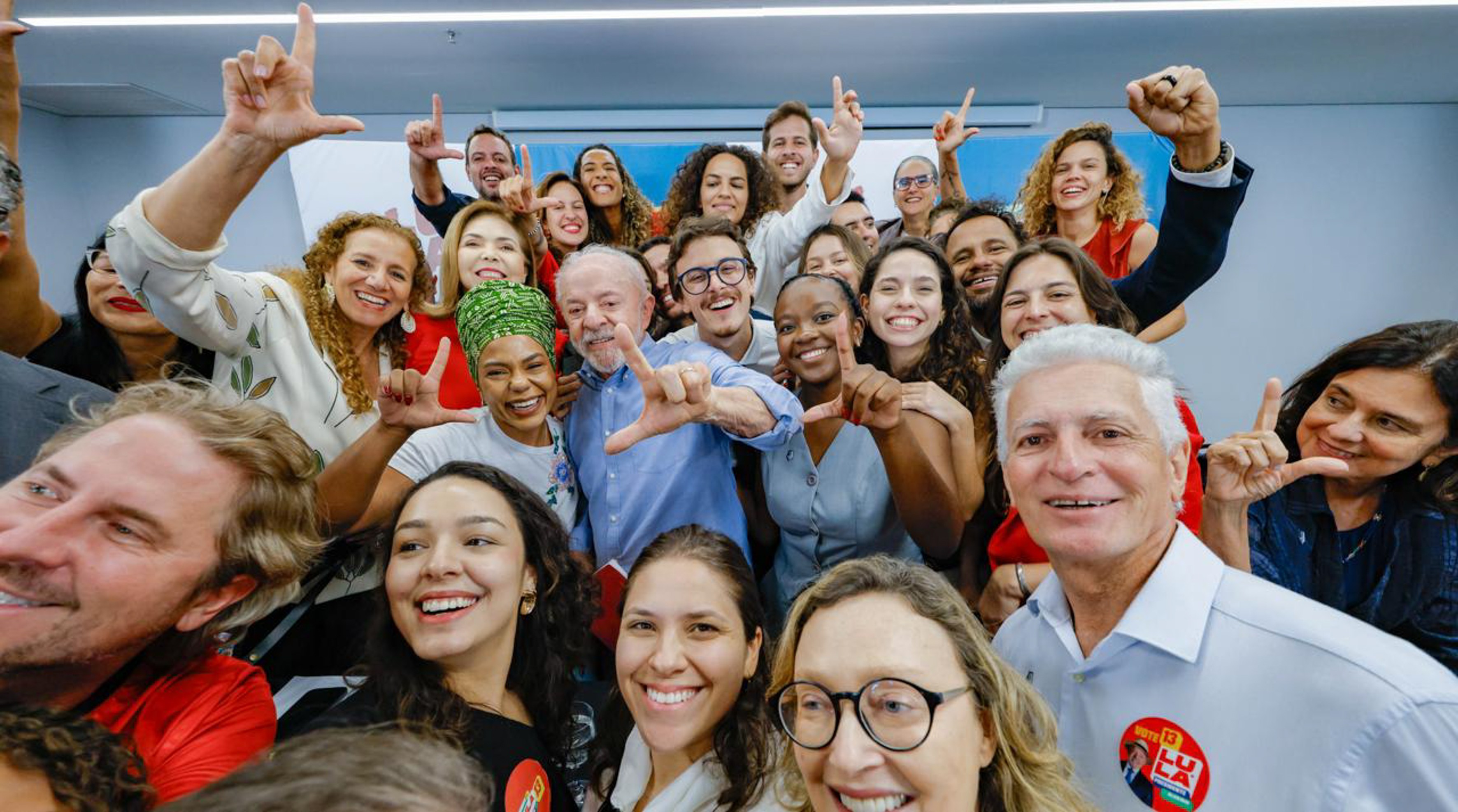 Um grupo diverso de pessoas sorrindo e fazendo o sinal de L com as mãos, incluindo o ex-presidente Lula no centro, vestindo camisa azul. Todos parecem felizes e engajados, em um ambiente interno claro