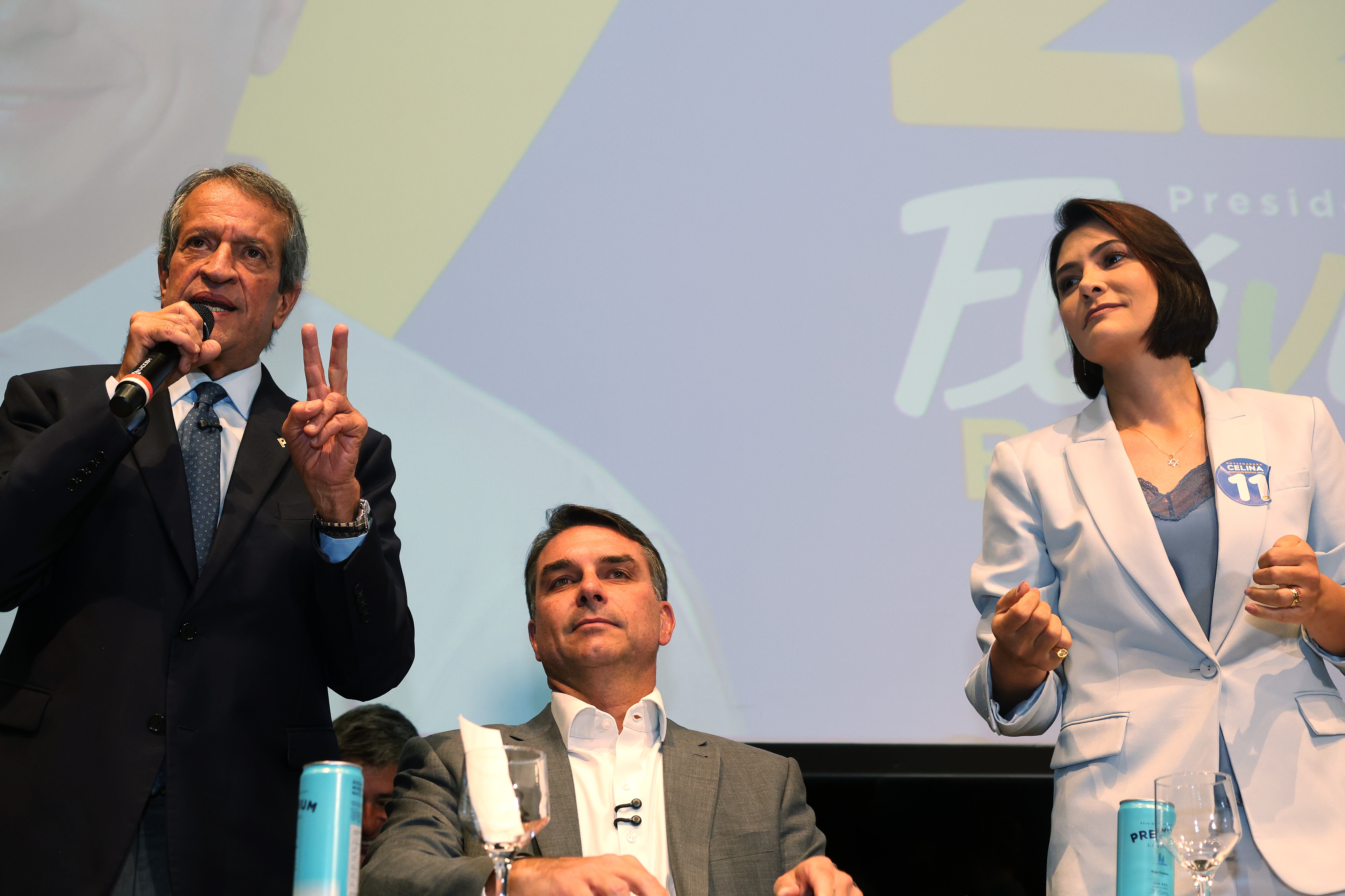 Um homem de terno escuro e gravata azul fala ao microfone, fazendo um sinal de vitória com a mão esquerda. Ao centro, outro homem de terno cinza e camisa branca está sentado, olhando para cima. À direita, uma mulher de blazer azul claro e blusa de renda azul escuro, com um broche 11 na lapela, olha para o lado esquerdo. Ao fundo, um telão com texto ilegível