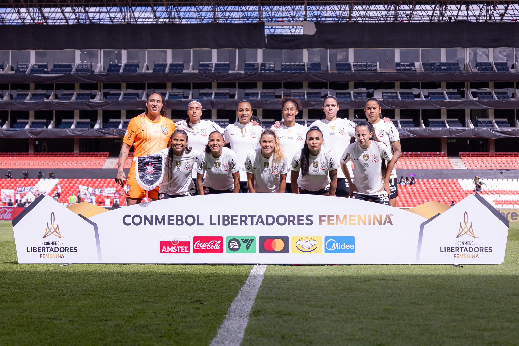 Time de futebol feminino posando para foto em campo de grama verde, com arquibancadas vazias ao fundo. Uma mulher de camisa laranja segura um escudo, enquanto as outras 11 usam uniformes brancos e sorriem. Um banner branco com CONMEBOL LIBERTADORES FEMENINA e logotipos de patrocinadores está à frente