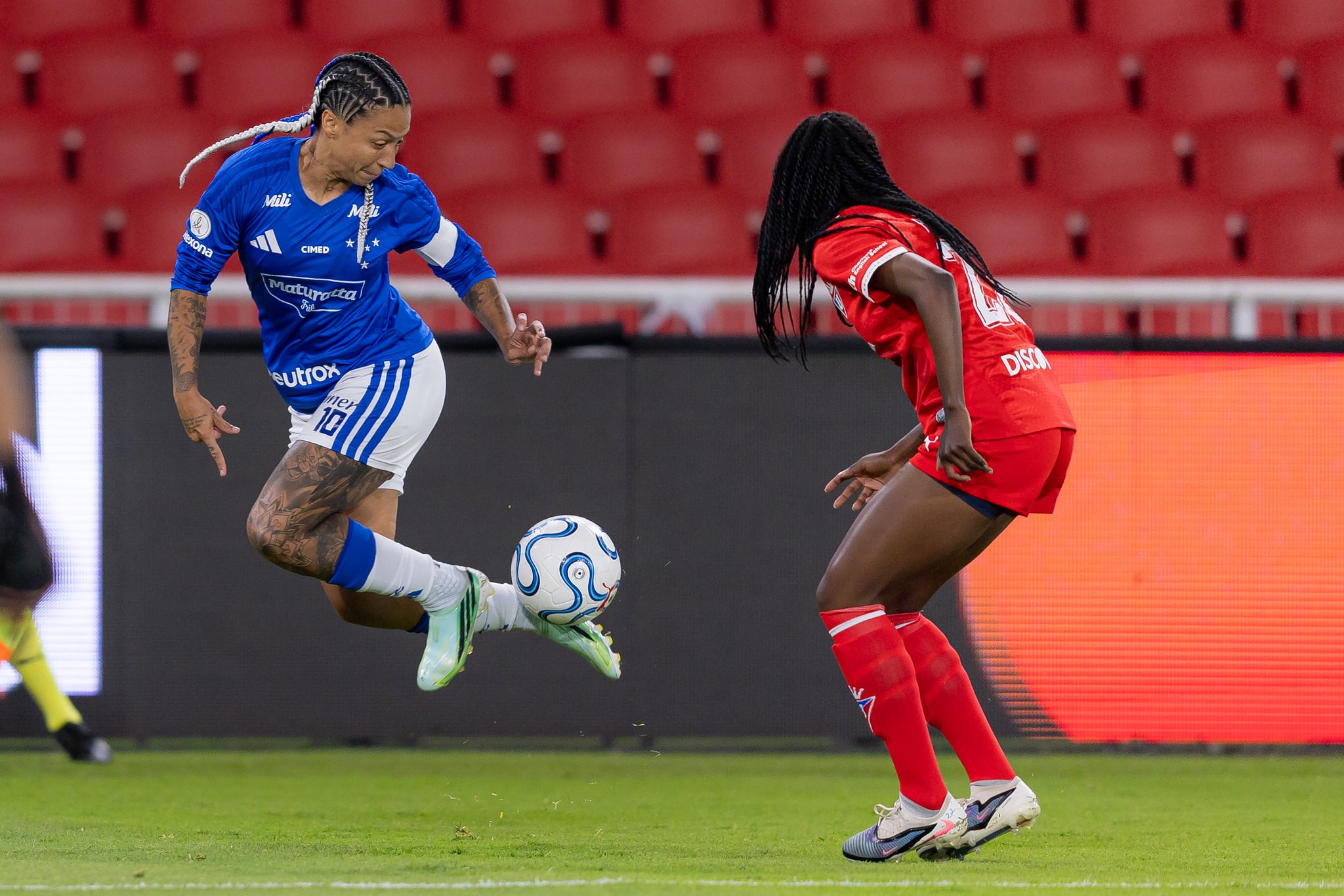 Duas jogadoras de futebol em campo. Uma mulher de tranças e uniforme azul e branco chuta a bola no ar, enquanto outra mulher de dreads e uniforme vermelho a observa, com arquibancadas vermelhas ao fundo