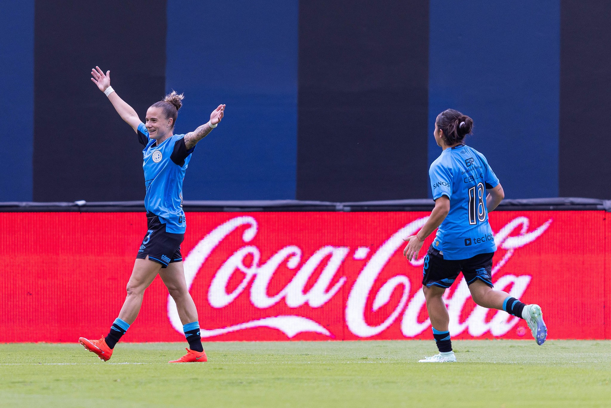 Duas jogadoras de futebol em campo, uma de costas com a camisa 18 e outra de frente com os braços erguidos em comemoração, ambas vestindo uniforme azul e preto. Ao fundo, um painel vermelho com o logo branco da Coca-Cola