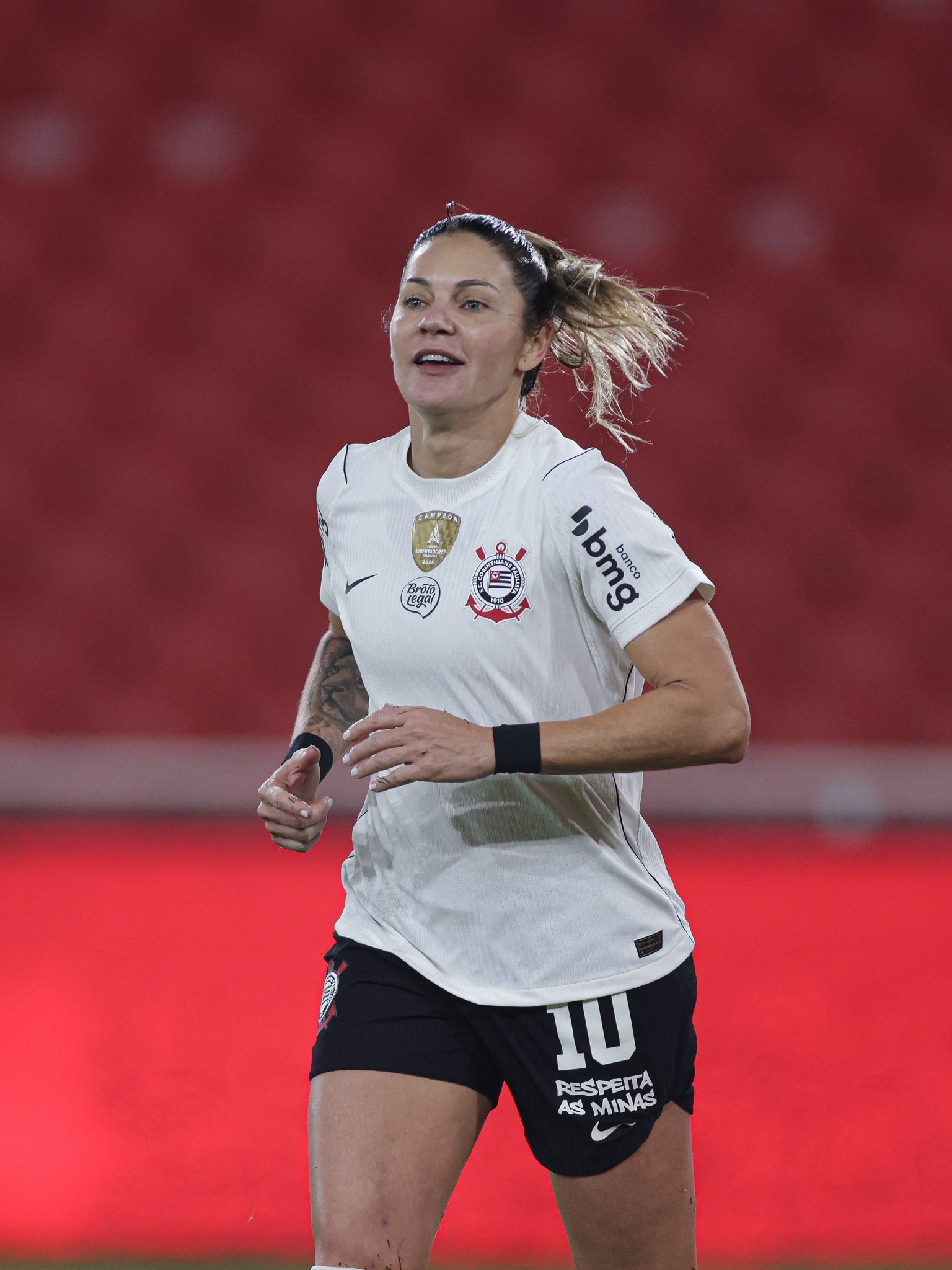 Jogadora de futebol feminina com cabelo preso, vestindo camisa branca do Corinthians com patrocínio Banco BMG e shorts pretos com a frase Respeita as Minas, correndo em campo com fundo vermelho desfocado