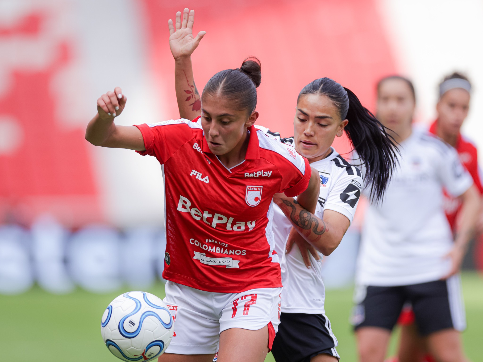 Duas jogadoras de futebol feminino em campo. Uma, de camisa vermelha e shorts brancos, domina a bola com o pé esquerdo, enquanto a outra, de camisa branca e shorts pretos, tenta marcá-la por trás. Ambas têm cabelos escuros presos e expressões concentradas.