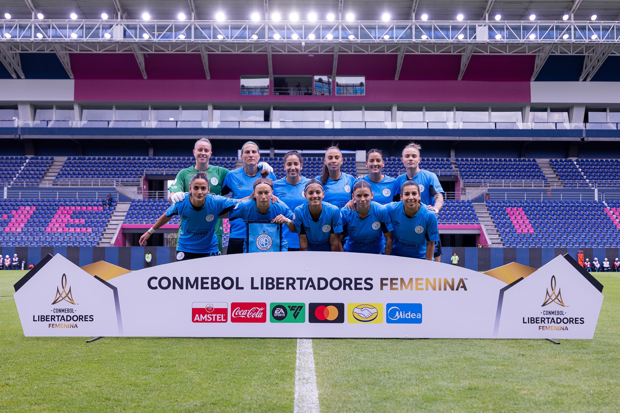 Time de futebol feminino posando para foto em campo de estádio, com arquibancadas azuis e roxas ao fundo. As 12 jogadoras vestem uniformes azuis e verdes, sorrindo. À frente, um banner branco com CONMEBOL LIBERTADORES FEMENINA e logotipos de patrocinadores.