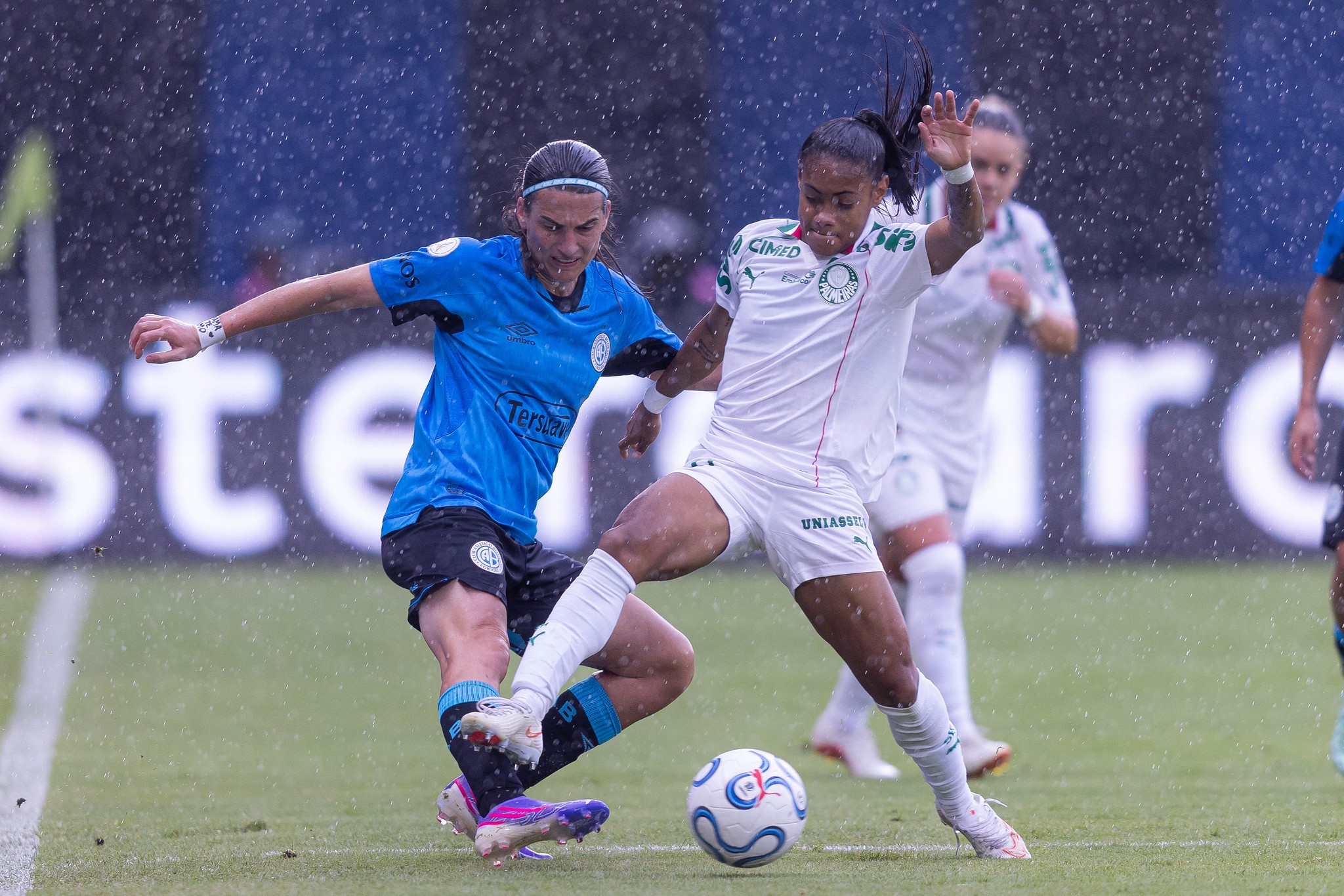 Duas jogadoras de futebol disputam a bola em campo molhado pela chuva. Uma, de camisa azul e shorts pretos, tenta driblar a outra, de camisa e shorts brancos, que estende a perna para interceptar a jogada. A bola está no chão, entre elas.