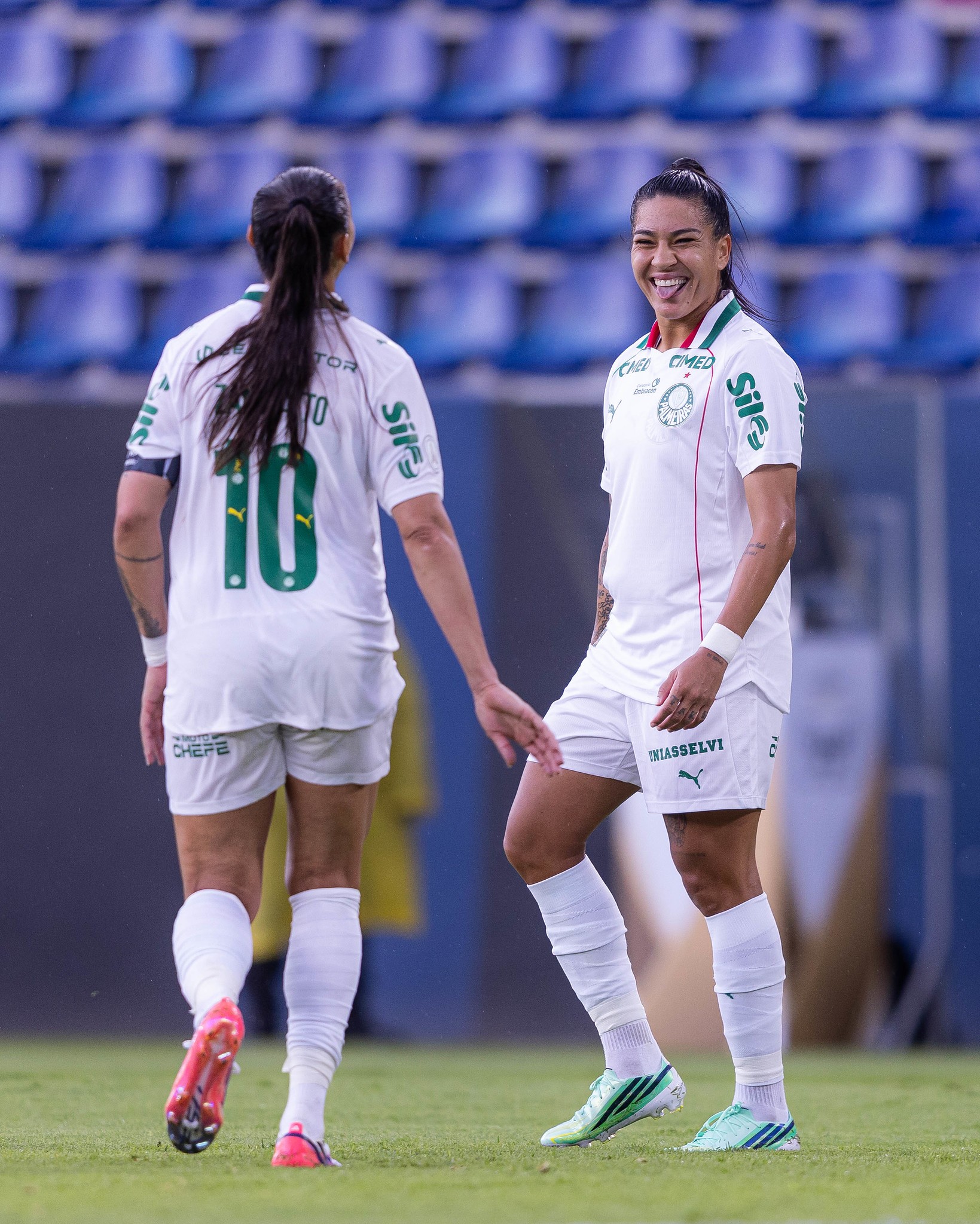 Duas jogadoras de futebol em campo. Uma de costas, com camisa 10, e outra sorrindo, com a língua para fora, vestindo uniforme branco e verde, em um estádio com cadeiras azuis ao fundo