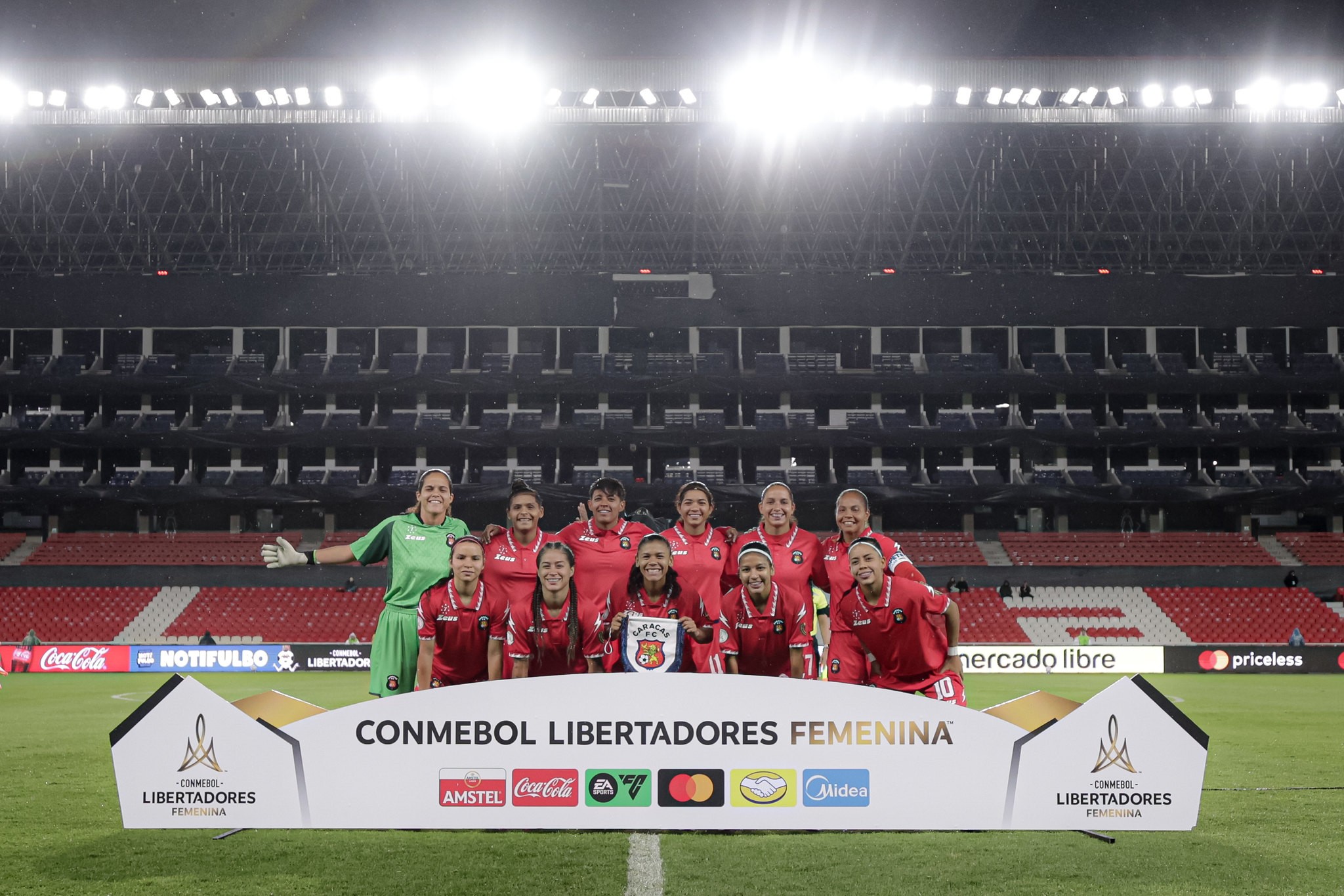 Time de futebol feminino posando para foto em campo de estádio à noite, com arquibancadas vazias e iluminação forte. Uma goleira de verde e dez jogadoras de vermelho sorriem atrás de um banner branco da CONMEBOL Libertadores Feminina