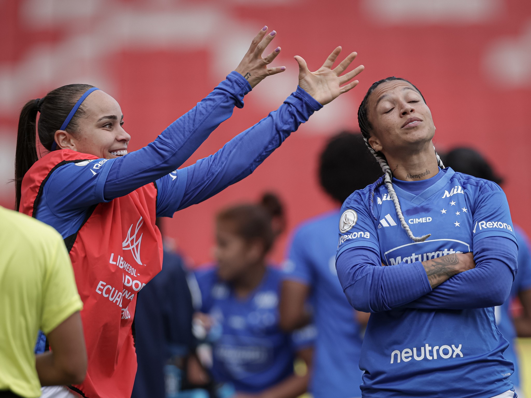 Duas jogadoras de futebol em campo. Uma mulher sorridente, de colete vermelho e camisa azul, gesticula com os braços erguidos. Ao lado, outra mulher de camisa azul, com tranças e braços cruzados, olha para cima com expressão de descontentamento. O fundo é vermelho e desfocado
