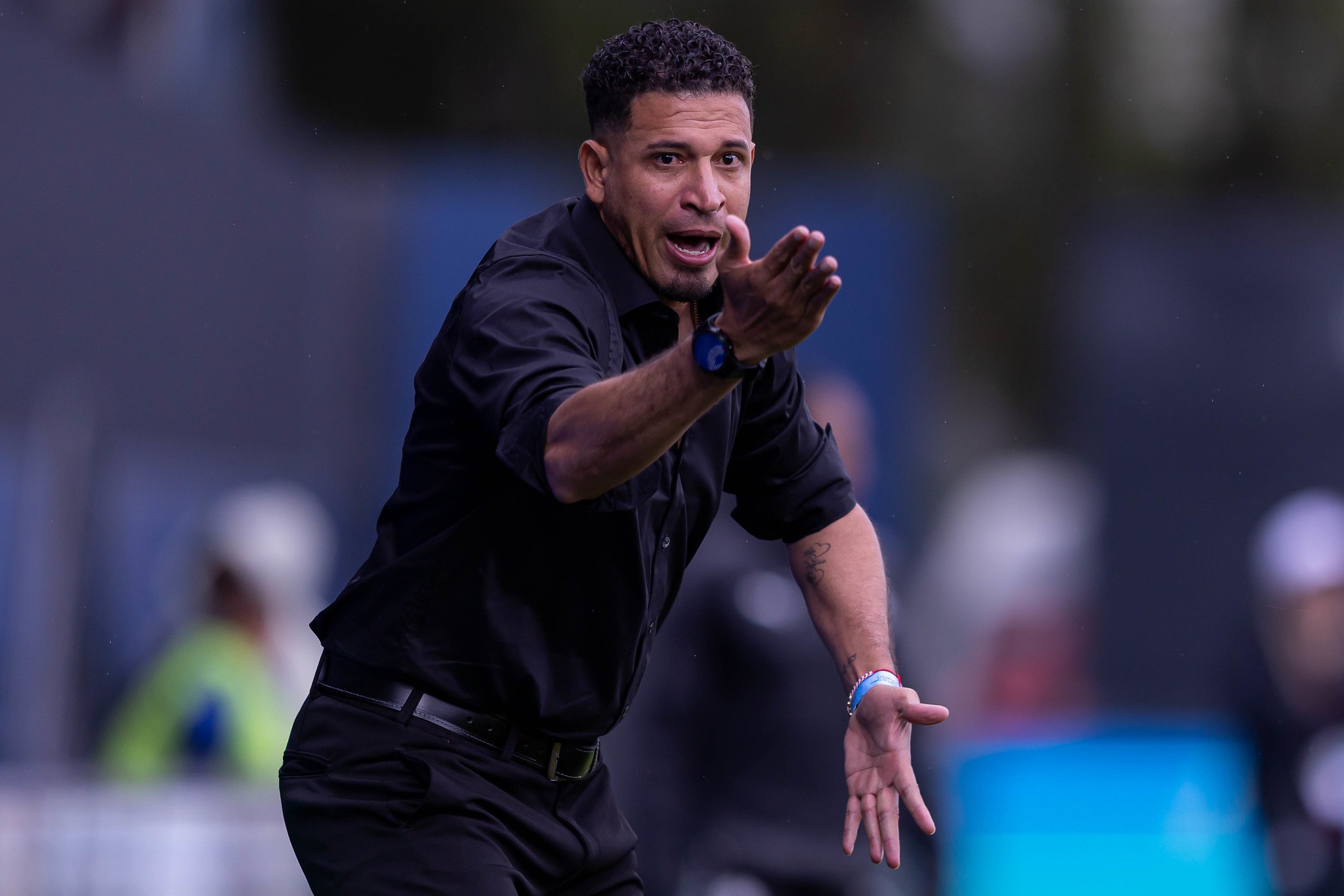 Homem de pele parda, cabelo crespo curto e barba rala, vestindo camisa social preta e calça preta, gesticula com a boca aberta e expressão séria, braços estendidos para frente, em um campo de futebol