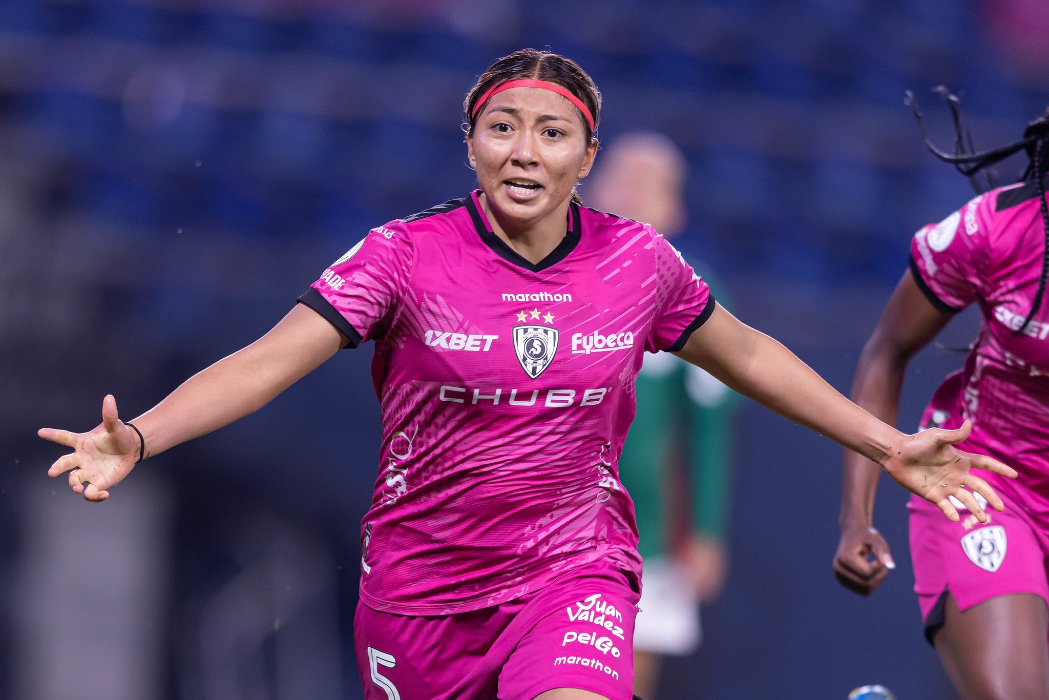 Jogadora de futebol com camisa e shorts rosa, faixa de cabelo vermelha, braços abertos e expressão de esforço em campo