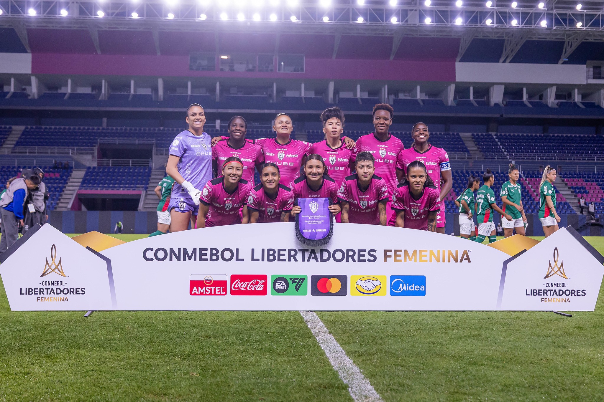 Time de futebol feminino com 11 jogadoras sorrindo, vestindo uniformes rosa escuro e roxo claro, posando em campo de grama verde à frente de um banner branco com CONMEBOL LIBERTADORES FEMENINA e logotipos de patrocinadores. Ao fundo, arquibancadas vazias de um estádio iluminado.