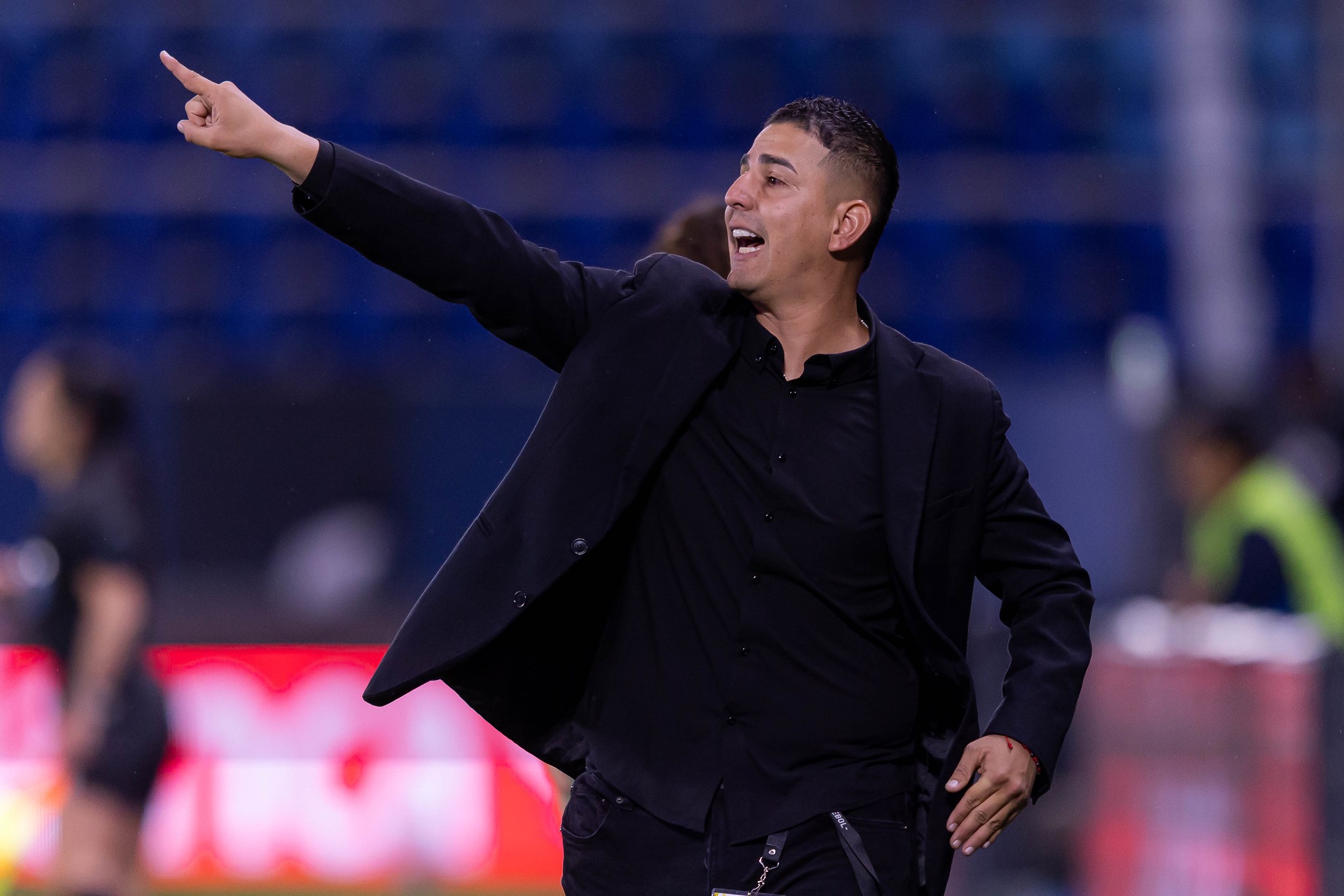 Homem de pele clara, cabelo curto e escuro, vestindo camisa e blazer pretos, aponta para cima com o braço direito estendido, boca aberta e expressão de comando em um campo de futebol