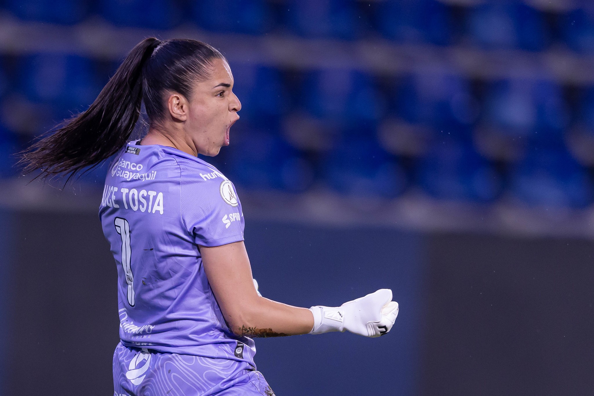 Goleira de cabelo preso, vestindo uniforme lilás com o nome KE TOSTA e número 1, gritando e comemorando em campo, usando luvas brancas, com arquibancada azul desfocada ao fundo