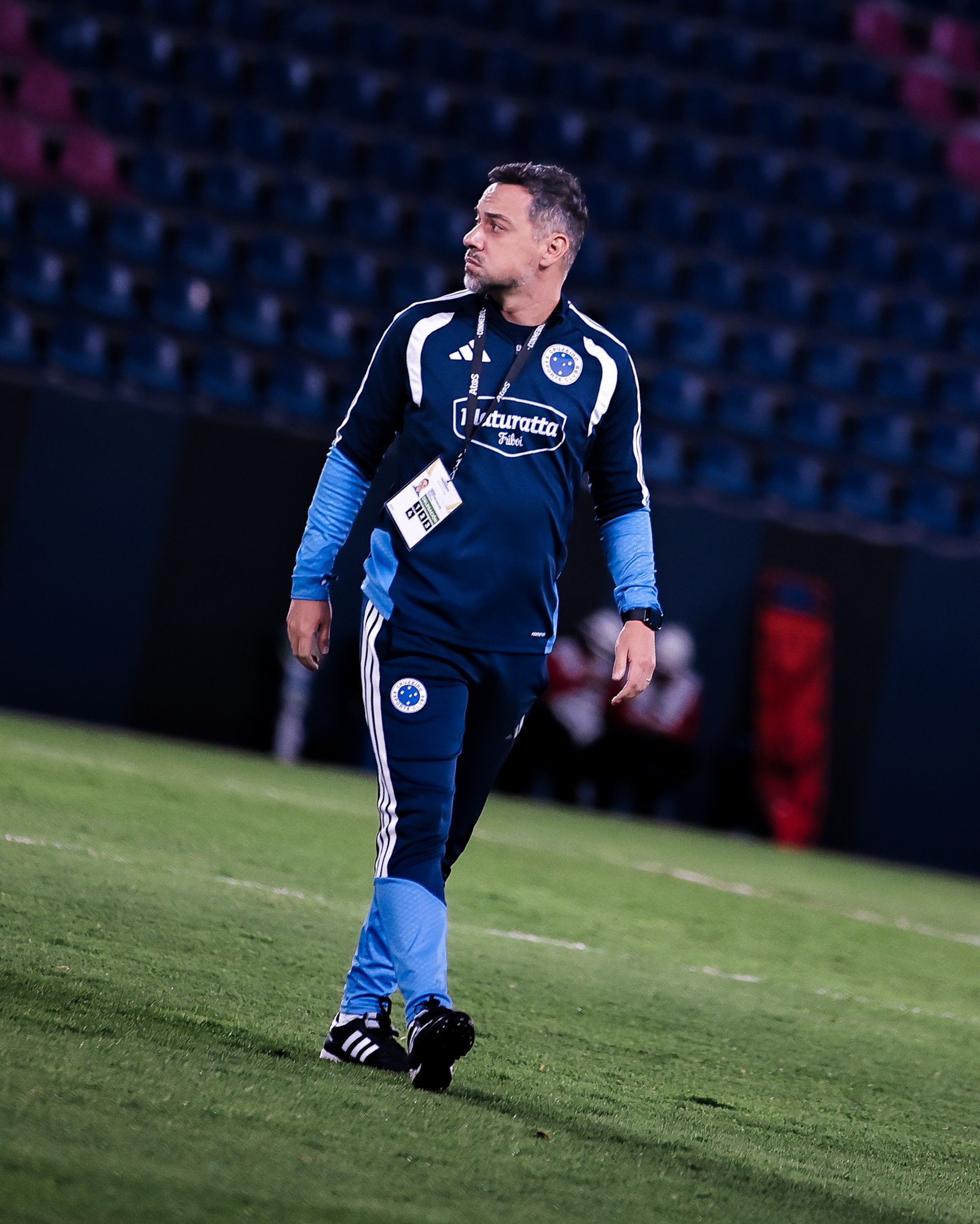 Homem de pele clara, cabelo curto e barba rala, vestindo agasalho azul-marinho e azul-claro com listras brancas, caminhando em campo de futebol gramado, olhando para a direita. Ao fundo, arquibancadas escuras.