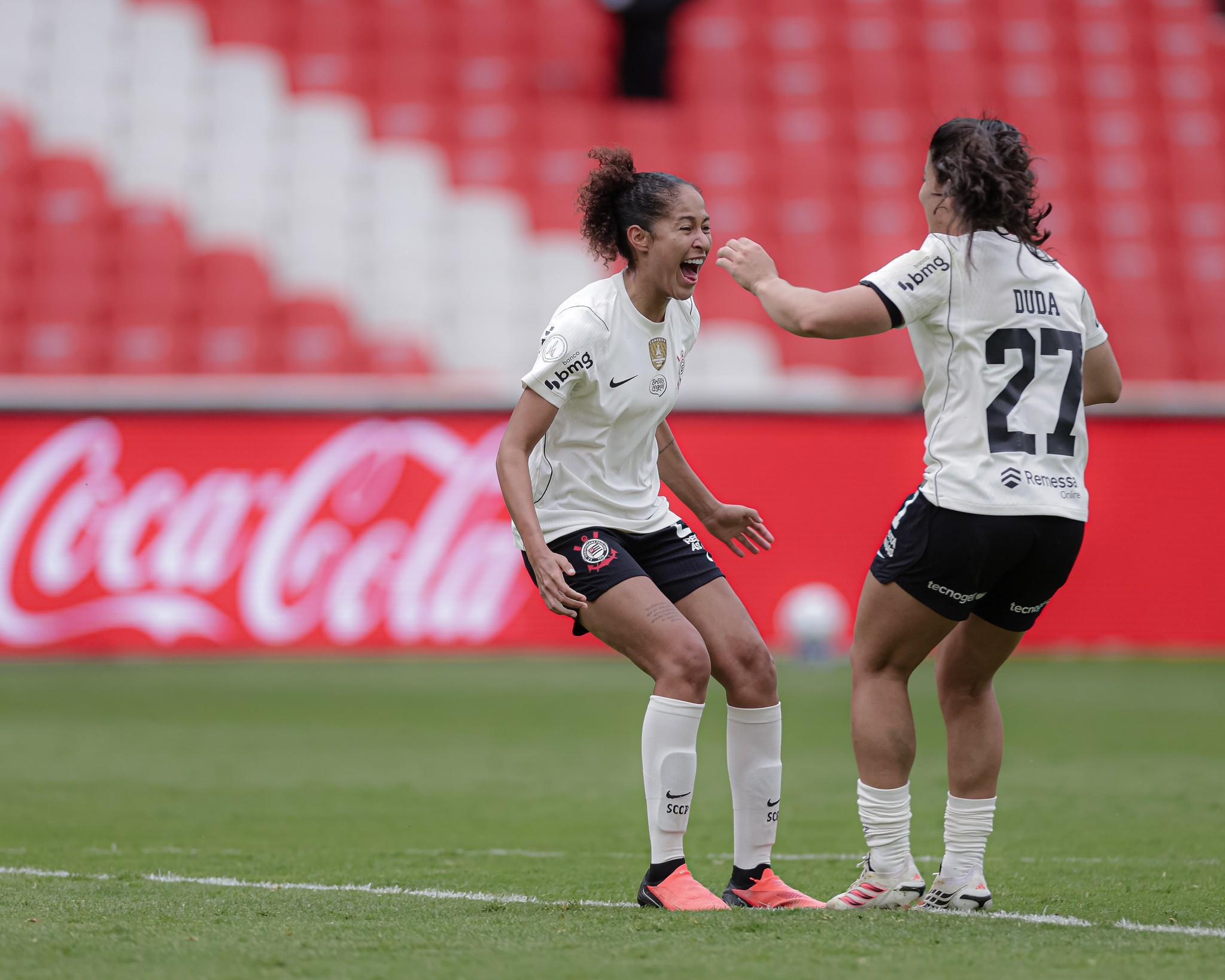 Duas jogadoras de futebol em campo, uma comemorando com a boca aberta e a outra tocando seu rosto, ambas vestindo uniformes brancos e pretos. Ao fundo, arquibancadas vermelhas e brancas e um letreiro da Coca-Cola