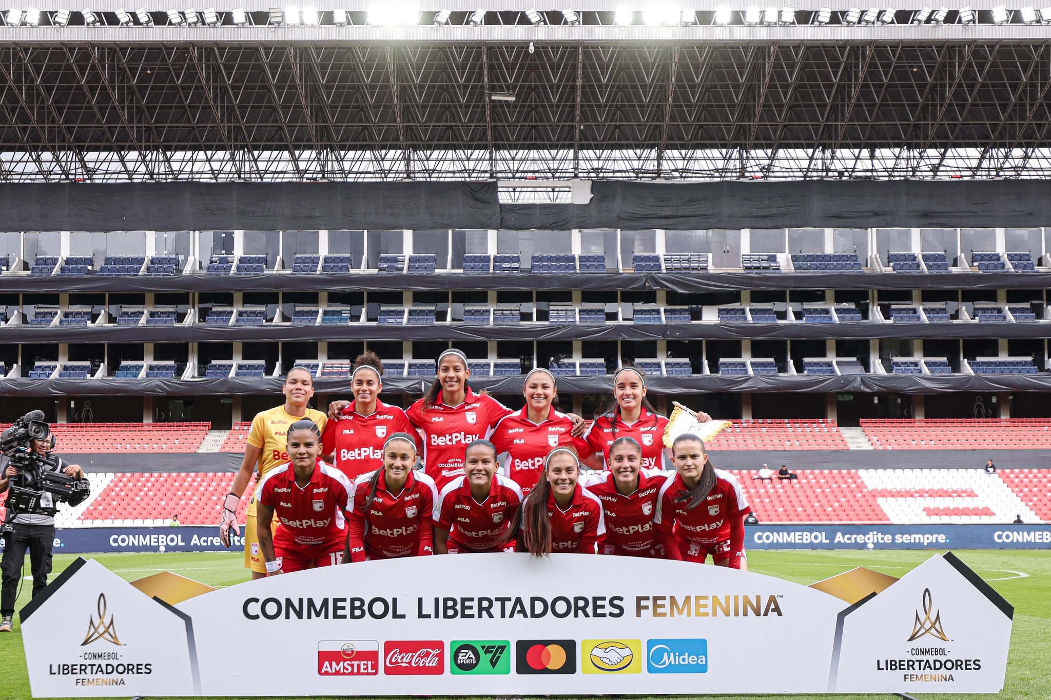 Time de futebol feminino posando para foto em campo de estádio vazio, com arquibancadas vermelhas e brancas ao fundo. As jogadoras vestem uniformes vermelhos e uma goleira usa amarelo. Elas sorriem, com um banner da Conmebol Libertadores Feminina à frente.