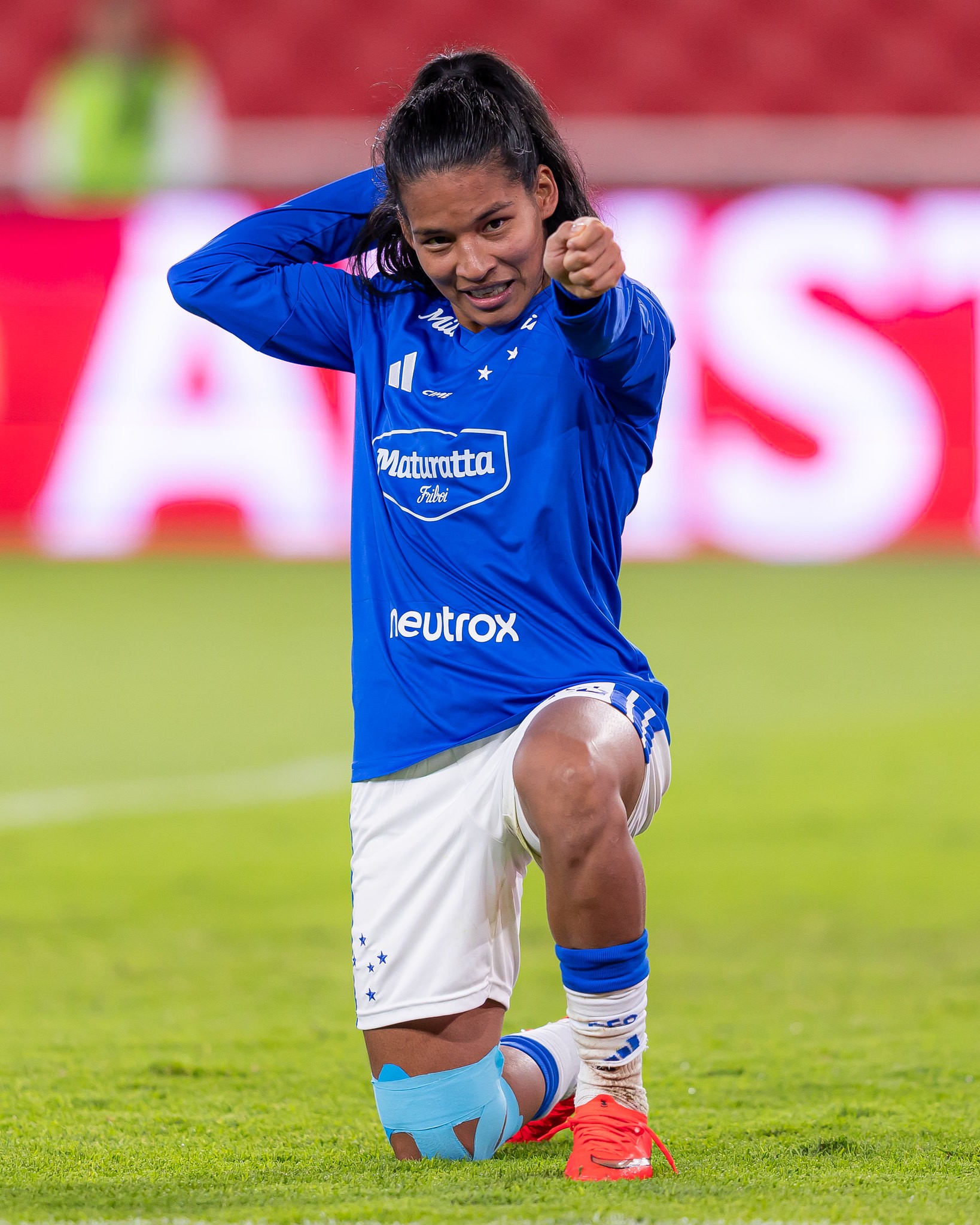 Jogadora de futebol com cabelo preso, vestindo camisa azul e shorts brancos, ajoelhada na grama, sorrindo e apontando para frente com o punho cerrado