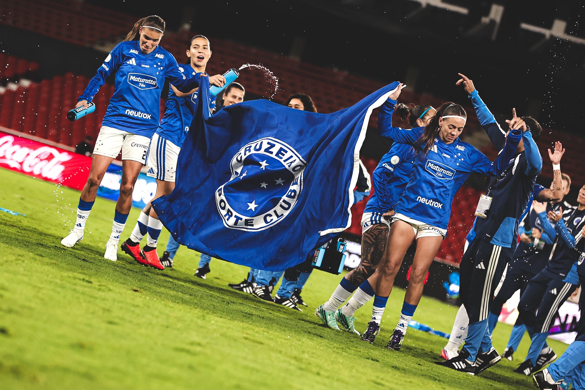 Jogadoras de futebol do Cruzeiro, vestindo uniformes azuis e brancos, comemoram em campo, uma delas segurando a bandeira do time e outra jogando água para cima. O gramado verde e as arquibancadas vermelhas do estádio são visíveis ao fundo, sob iluminação noturna