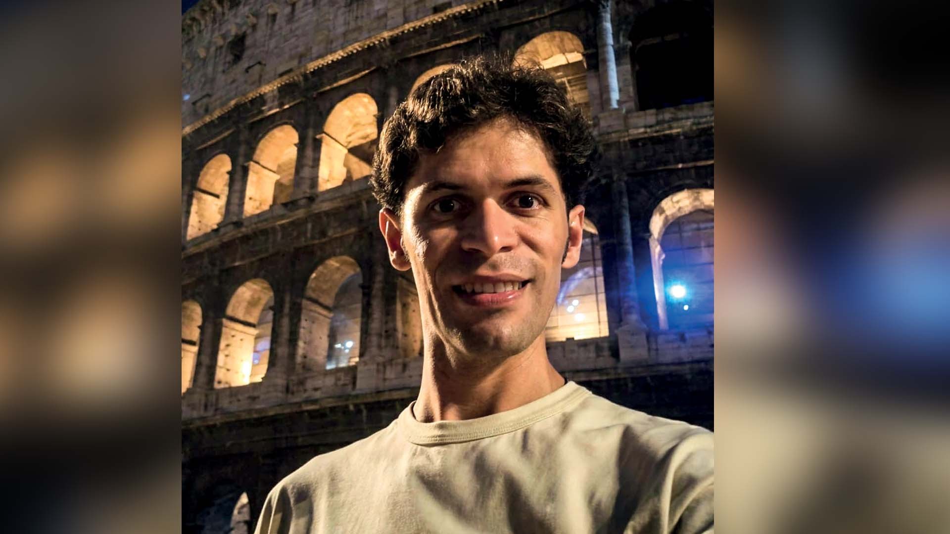 Homem de pele clara e cabelo escuro, sorrindo, com o Coliseu iluminado ao fundo, à noite