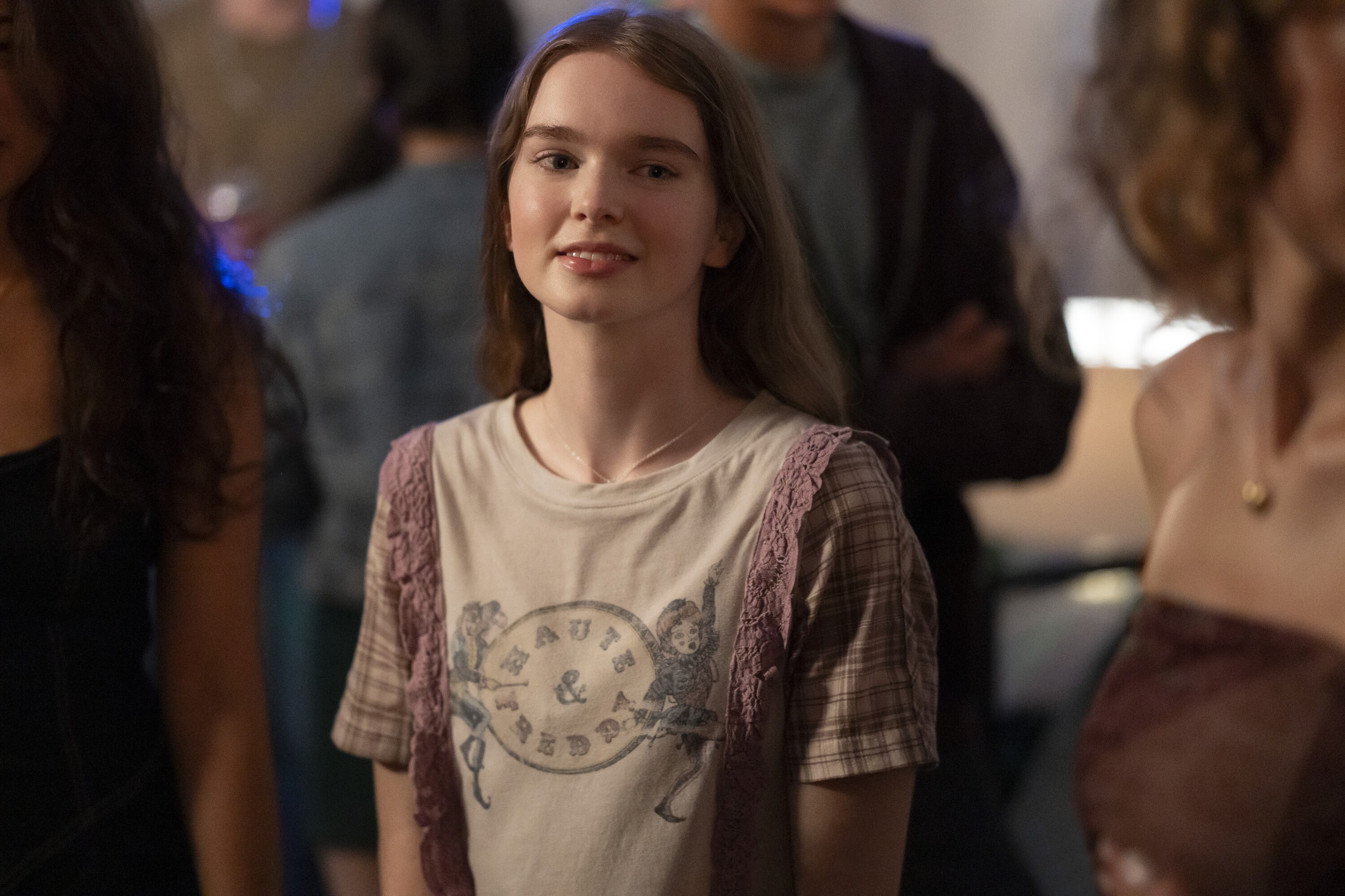Jovem de cabelos castanhos claros e lisos, sorrindo levemente, usando uma camiseta bege com estampa vintage e mangas xadrez marrom com detalhes de renda lilás, em um ambiente interno com pessoas desfocadas ao fundo