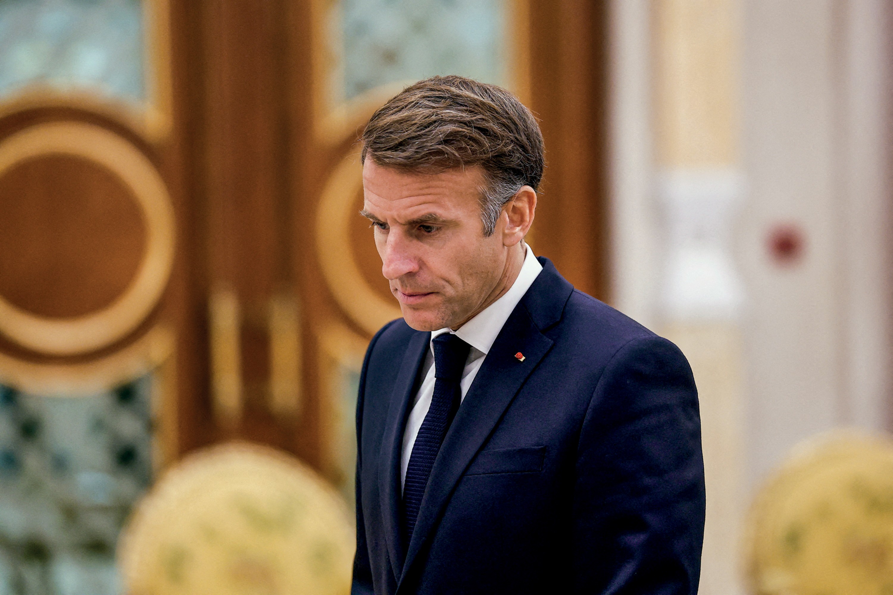 Emmanuel Macron, presidente francês, em close-up, olhando para baixo com expressão séria, vestindo terno azul-marinho e gravata escura, com um broche de bandeira francesa na lapela. O fundo está desfocado, com detalhes em madeira e dourado.