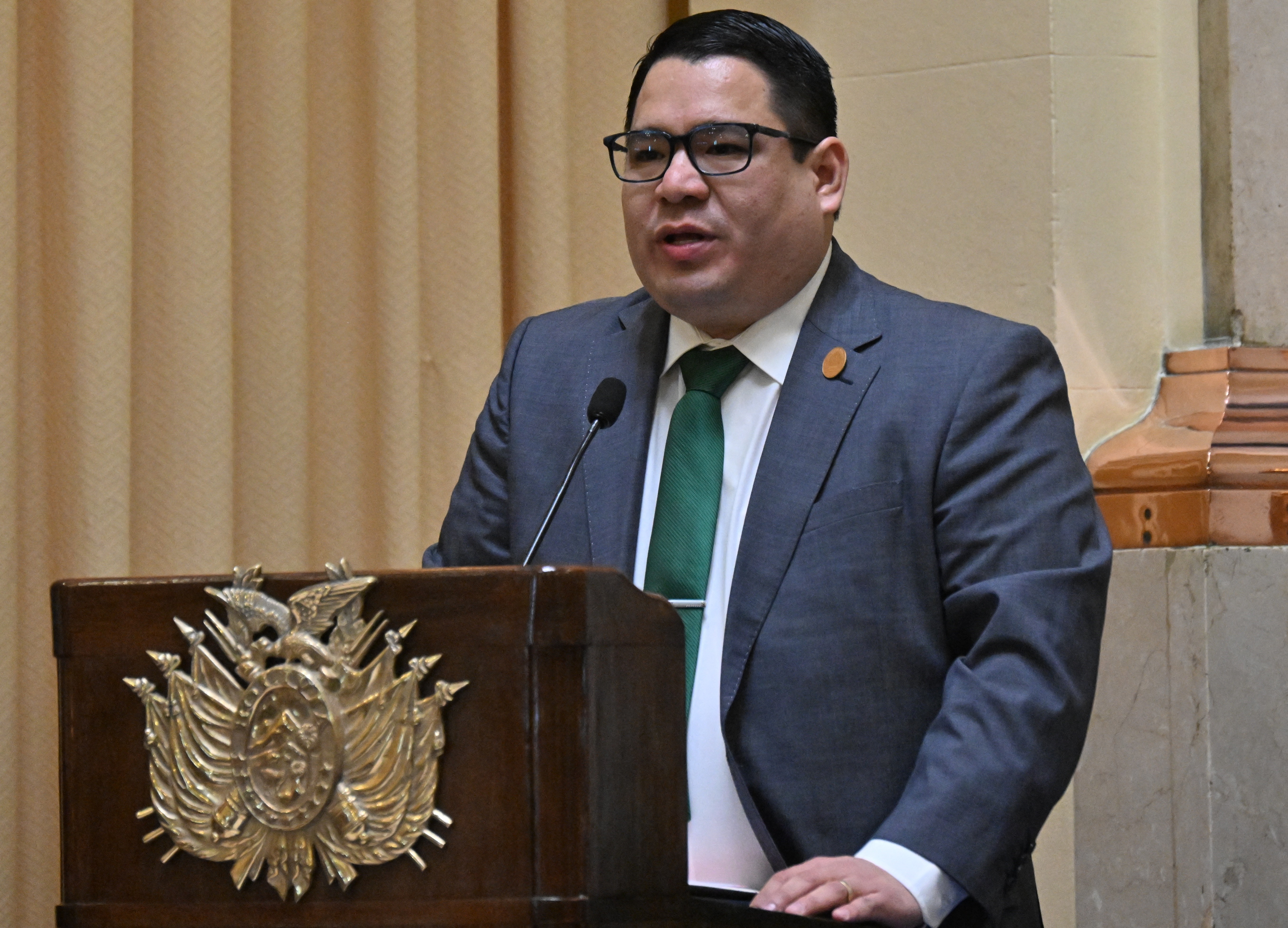 Homem de óculos, terno cinza e gravata verde, falando em um púlpito de madeira com um brasão dourado, em um ambiente interno com cortinas bege ao fundo