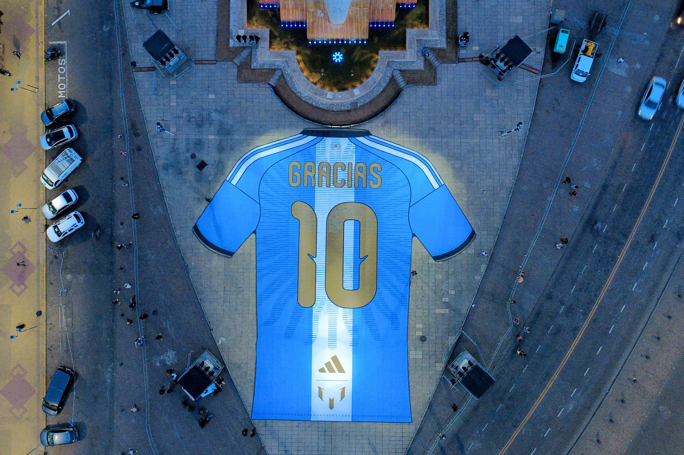 Vista aérea de uma camisa gigante da seleção argentina, azul e branca, com o número 10 e a palavra GRACIAS em dourado, iluminada no centro de uma praça com carros e pessoas ao redor