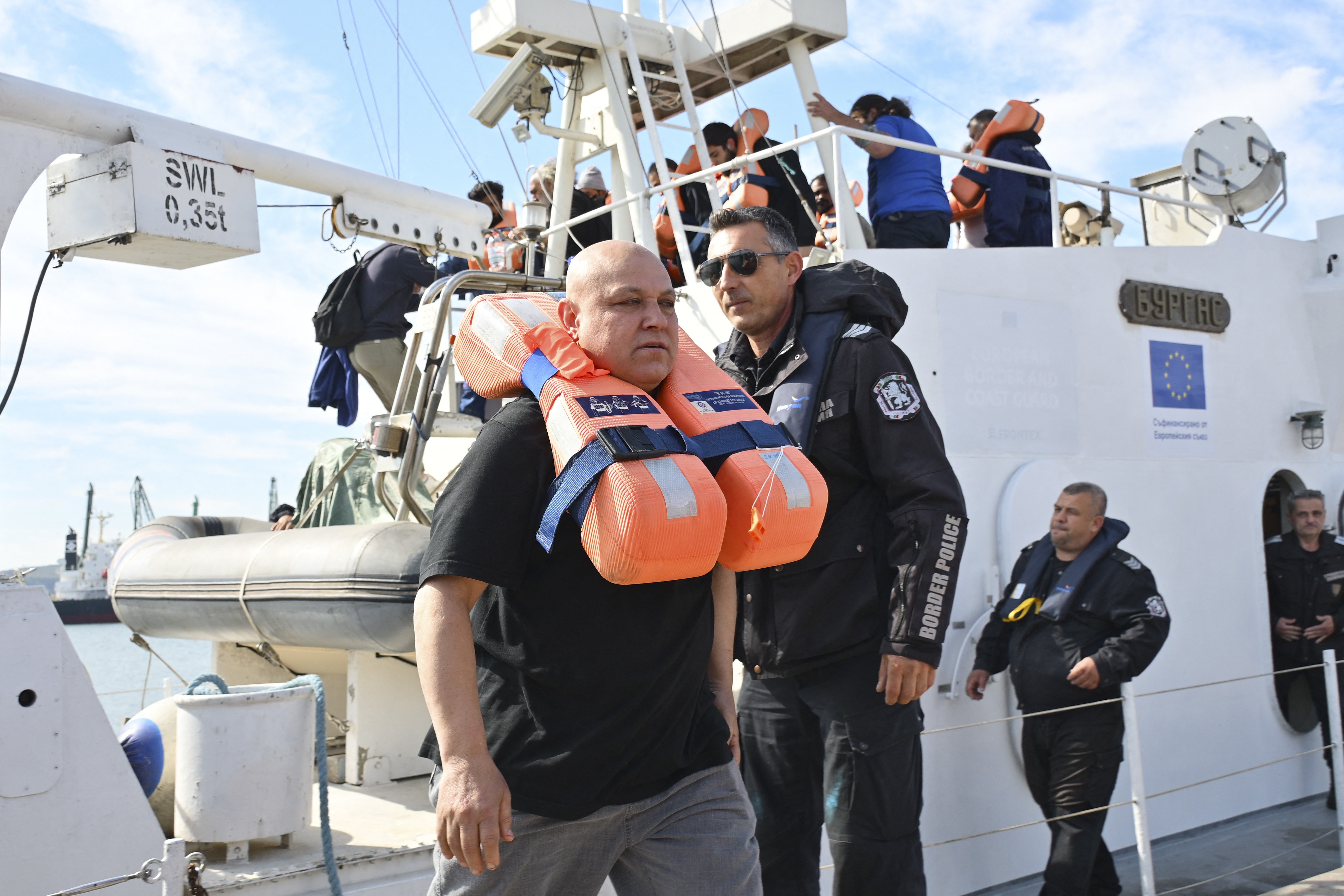 Homem careca com colete salva-vidas laranja no pescoço, acompanhado por um policial de óculos escuros e uniforme Border Police, em um barco branco com a bandeira da União Europeia. Outras pessoas estão no convés, algumas com coletes, e um bote inflável está amarrado ao lado. O céu é azul com nuvens esparsas