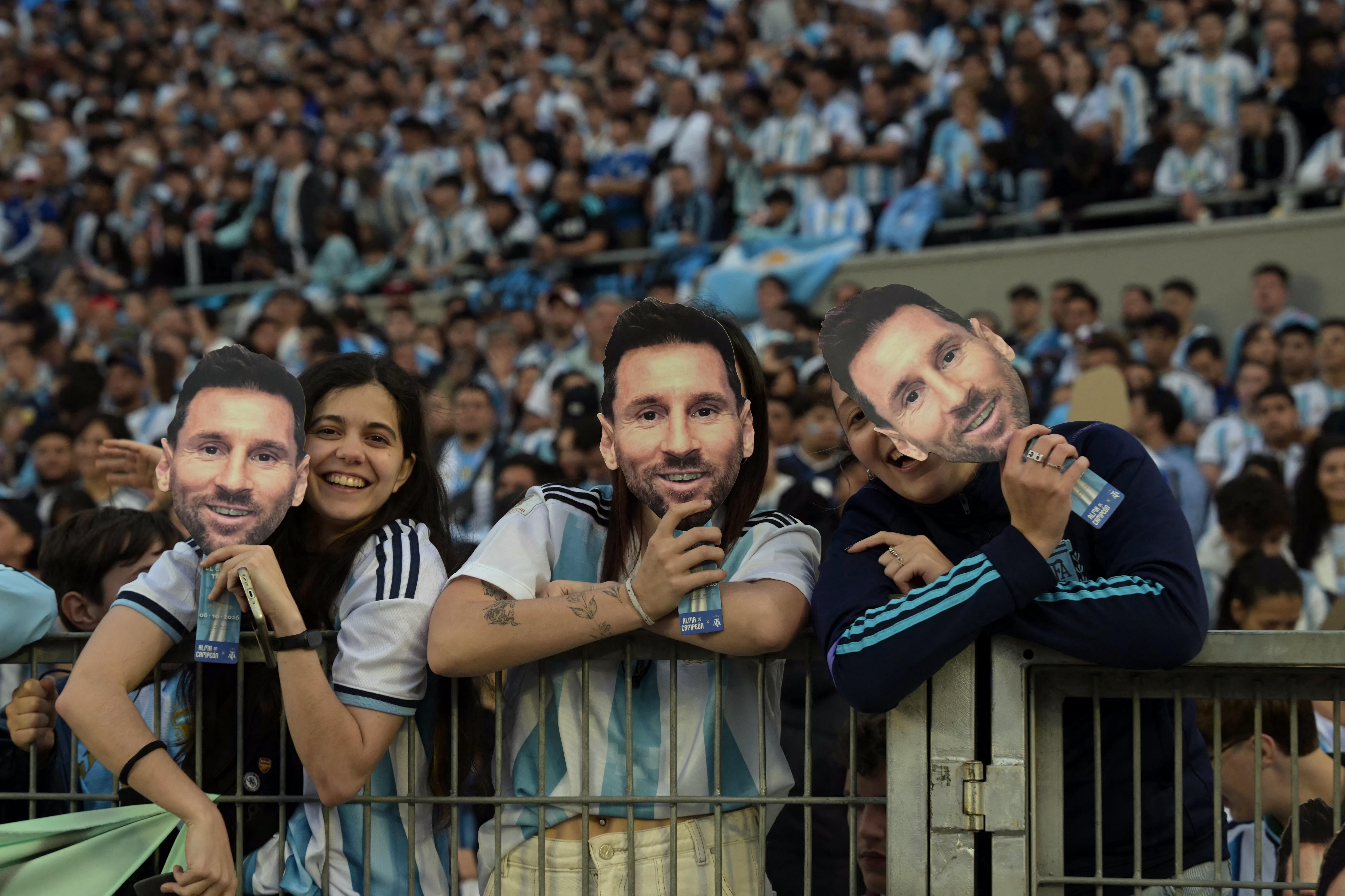 Torcedores argentinos, incluindo uma mulher sorridente e duas pessoas segurando máscaras do rosto de Messi, assistem a um jogo no estádio, vestindo camisas celestes e brancas, com a arquibancada lotada ao fundo