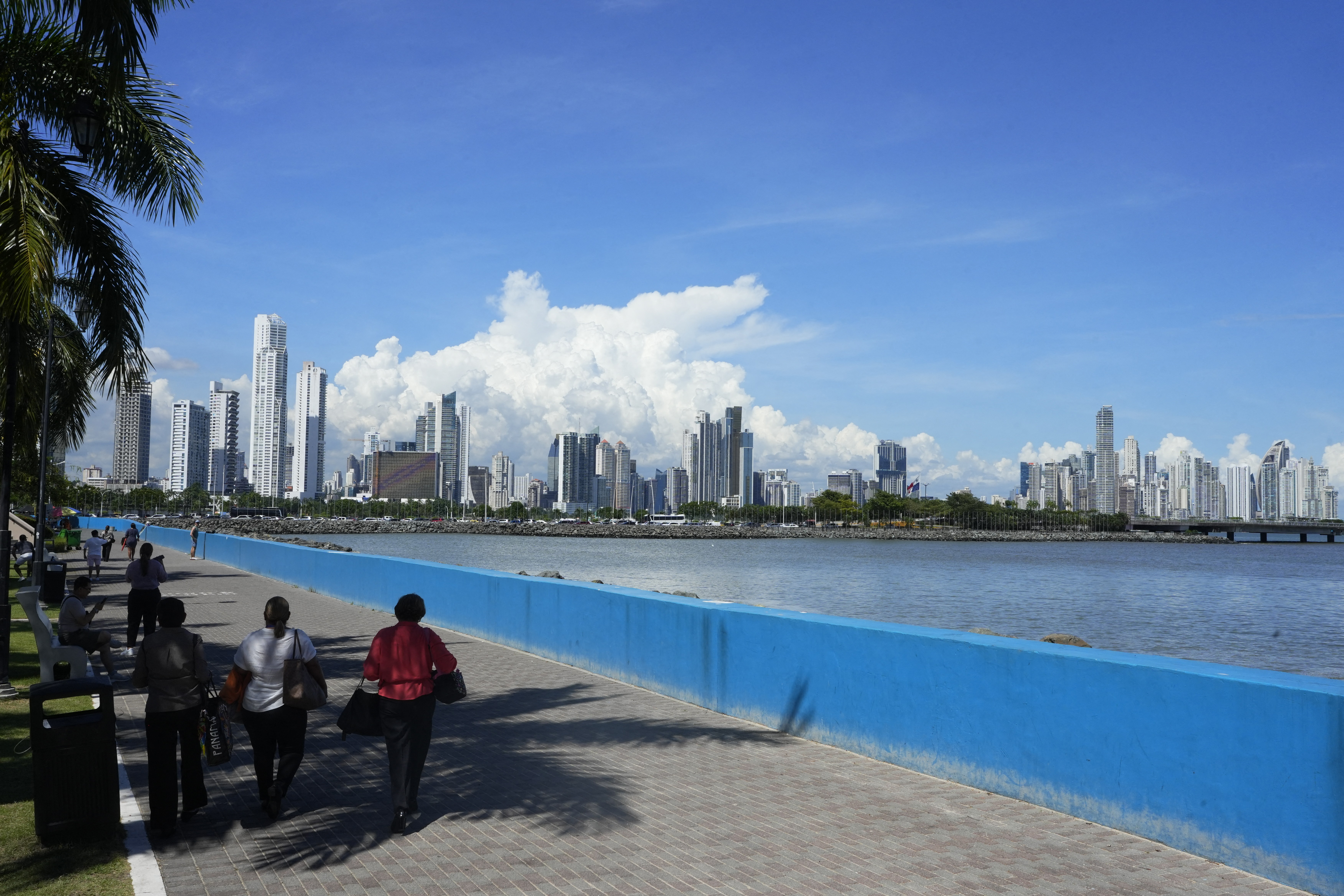 Pessoas caminham em calçadão de pedra cinza com um muro azul-claro à direita, margeando um rio. Ao fundo, uma cidade com arranha-céus brancos e cinzas sob céu azul com nuvens brancas. Palmeiras à esquerda projetam sombras no chão