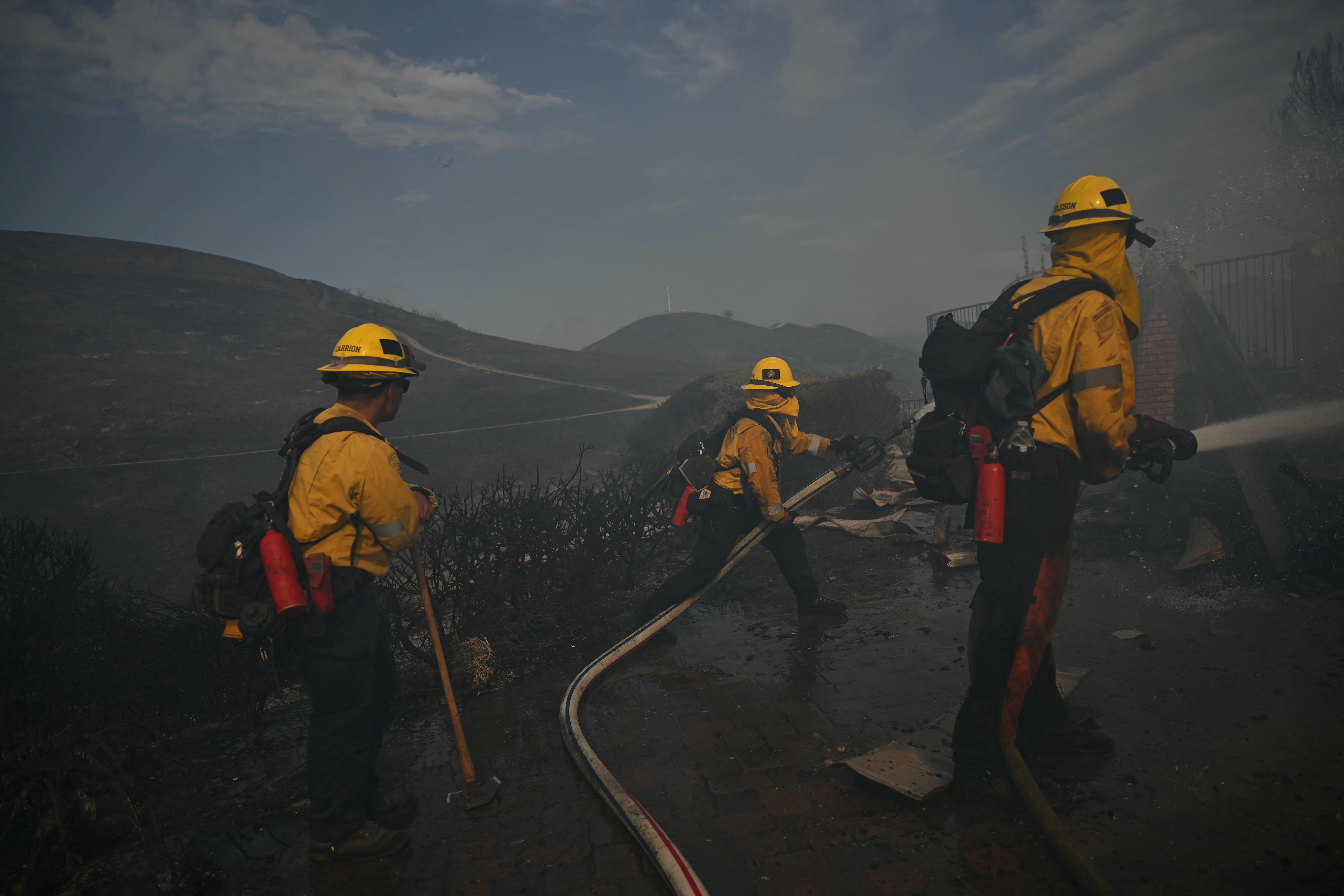 Três bombeiros com capacetes e uniformes amarelos trabalham em um cenário de fumaça e destruição. Um deles, à direita, joga água com uma mangueira. Outro, ao centro, segura uma mangueira no chão. O terceiro, à esquerda, observa com uma ferramenta. O fundo mostra colinas escuras e um céu nublado, com uma estrada sinuosa. O chão está molhado e escuro, com detritos espalhados. A atmosfera é sombria e urgente, refletindo o combate a um incêndio florestal.