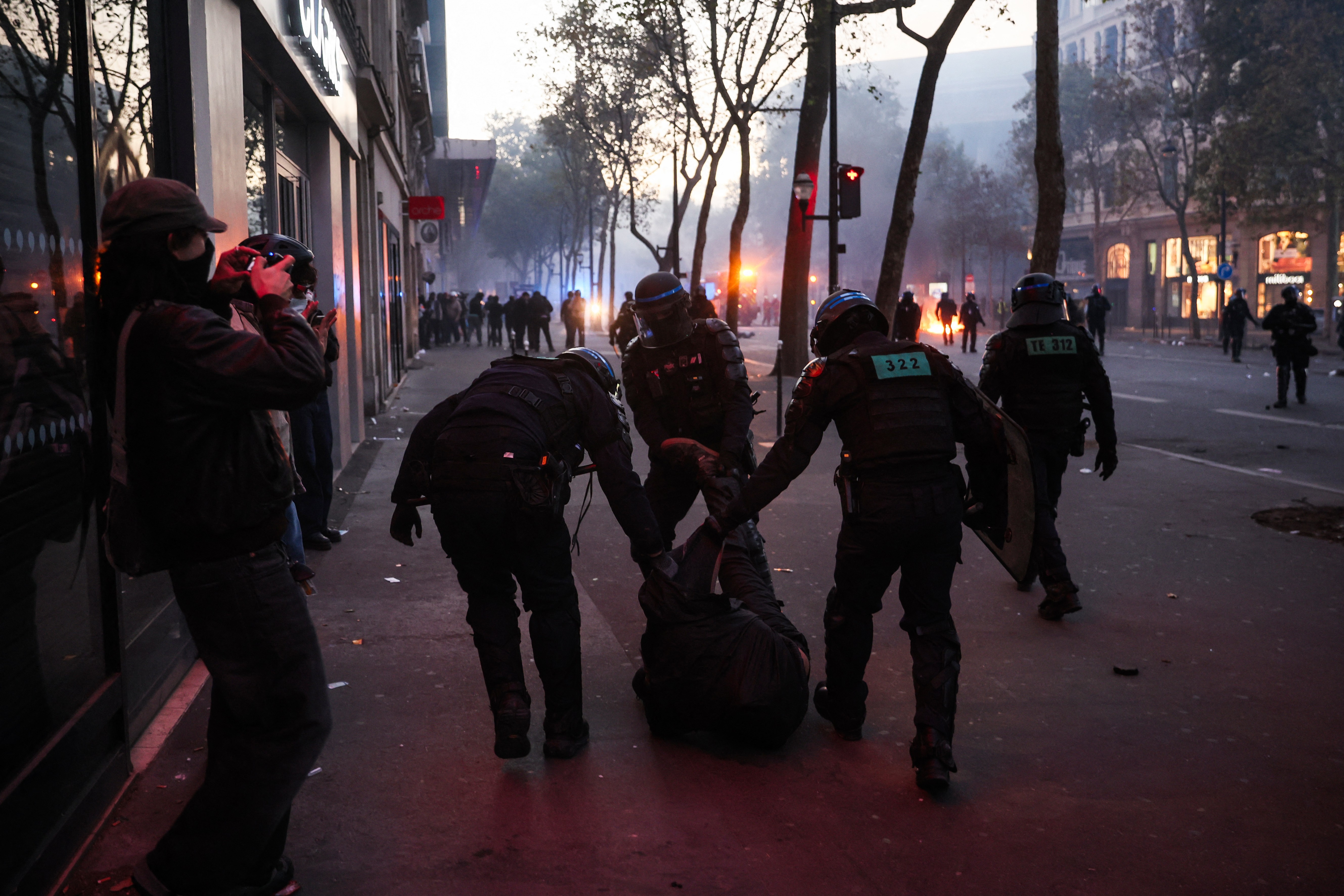 Três policiais com capacetes e coletes de proteção arrastam uma pessoa caída no chão, em uma rua urbana escura. À esquerda, um fotógrafo com boné e máscara facial registra a cena. Ao fundo, luzes de veículos e edifícios iluminam a névoa, com árvores sem folhas e mais pessoas distantes. O chão está sujo com detritos.