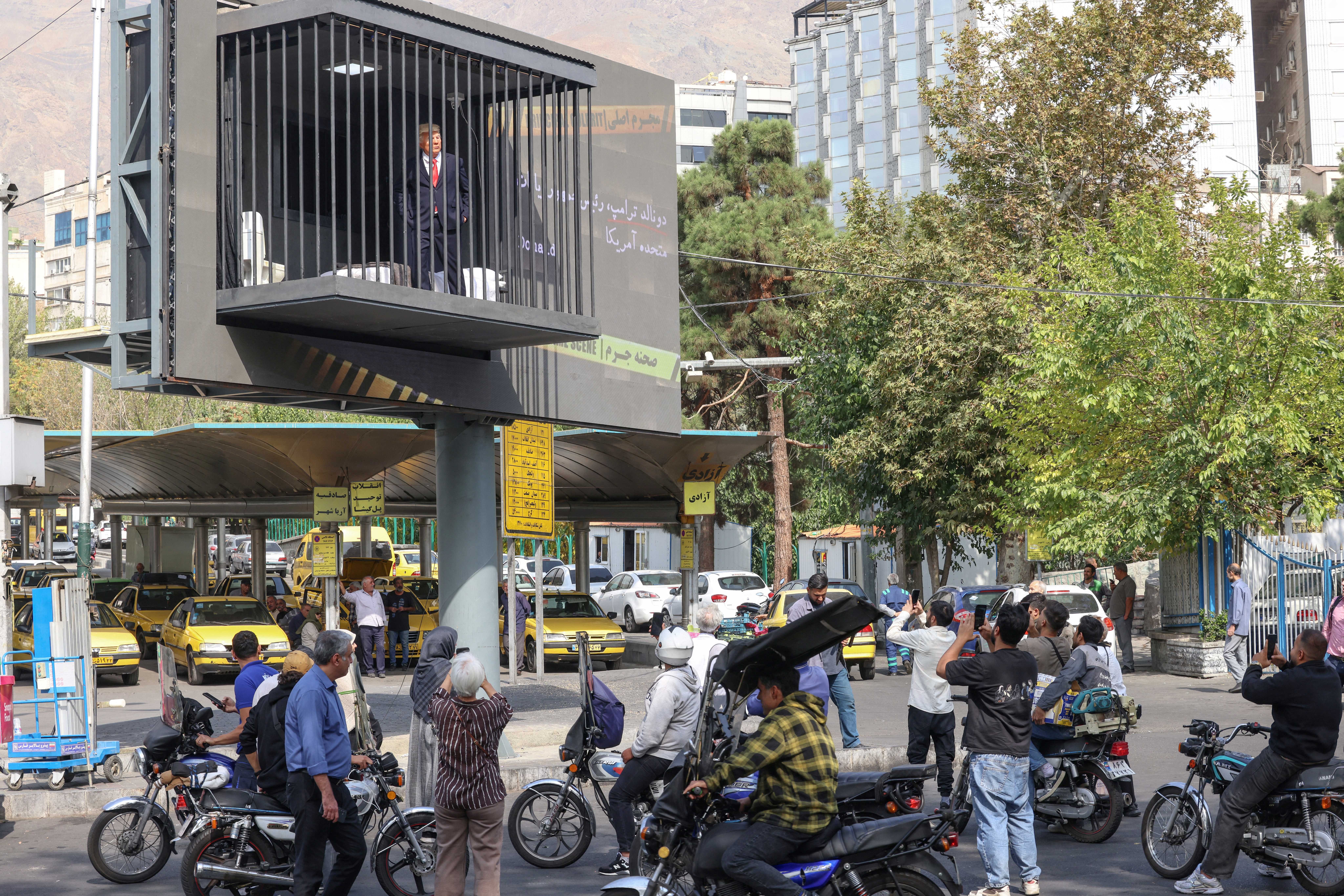 Um outdoor exibe uma figura masculina em terno e gravata, dentro de uma estrutura que simula uma jaula, sobre uma rua movimentada com pessoas e motocicletas. Ao fundo, prédios e árvores complementam a cena urbana