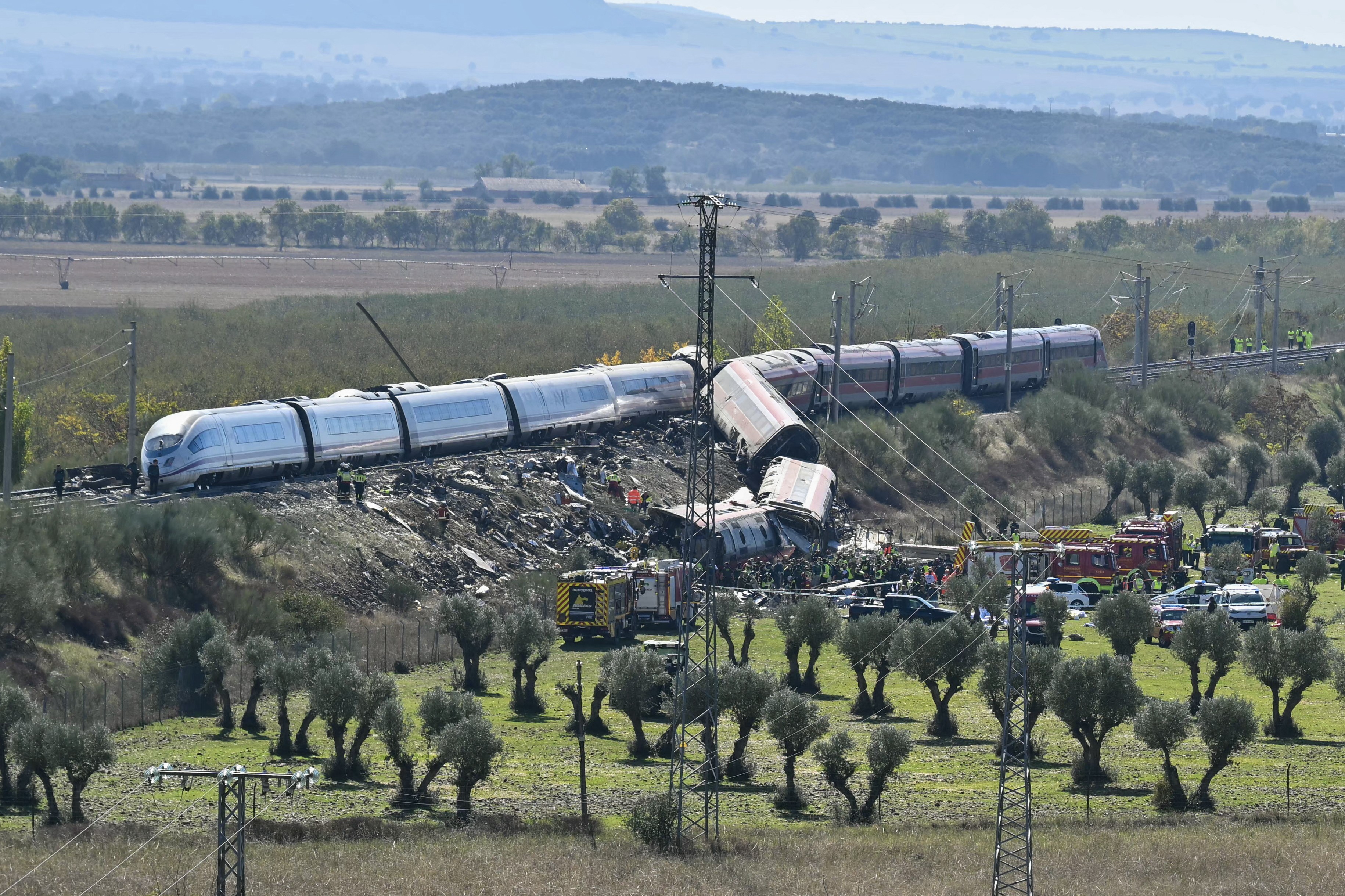 Trem descarrilado em área rural, com vagões tombados e equipes de resgate, incluindo bombeiros e veículos de emergência, trabalhando no local. A paisagem tem colinas, árvores e vegetação rasteira.