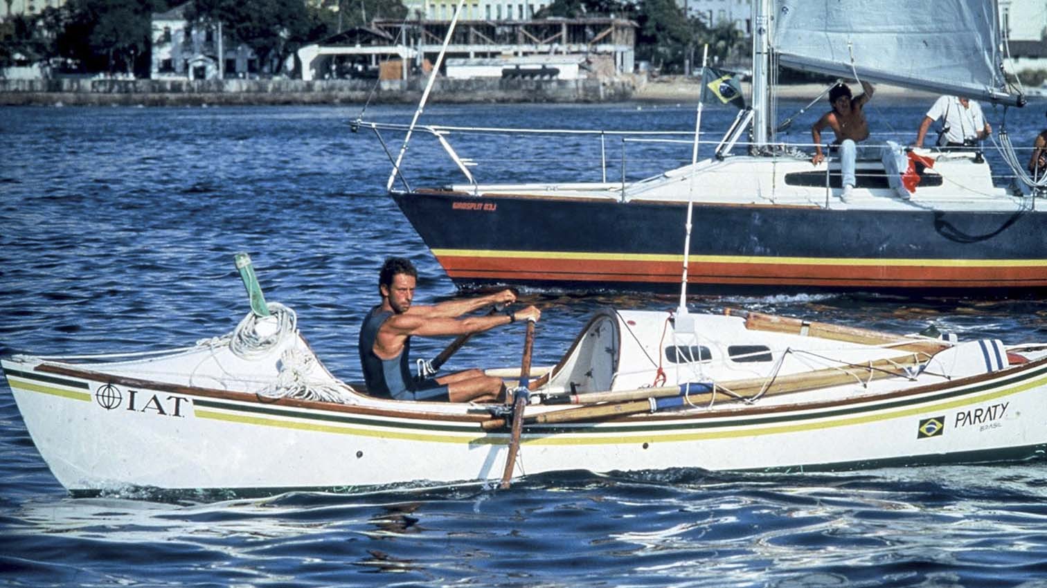 Amyr Klink remando vigorosamente em seu barco a remo branco IAT PARATY com detalhes verde e amarelo, em águas azuis. Ao fundo, um veleiro com a bandeira do Brasil e pessoas a bordo, e a orla de uma cidade