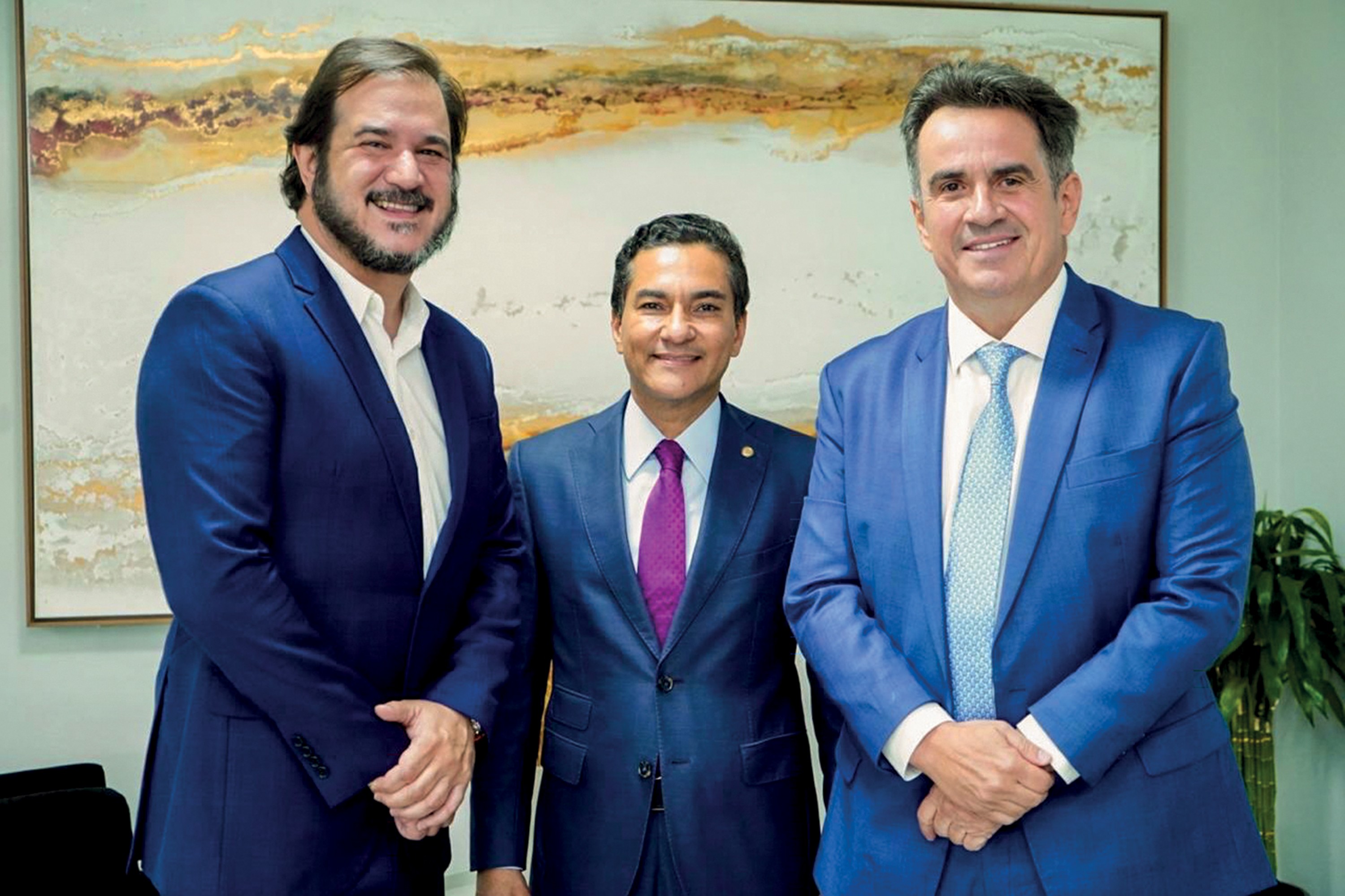Três homens sorridentes, de meia-idade, vestindo ternos azuis e camisas sociais, posam em pé. O homem da esquerda tem barba e camisa branca, o do centro usa gravata roxa e o da direita, gravata azul clara. Ao fundo, uma pintura abstrata em tons terrosos e uma planta verde.