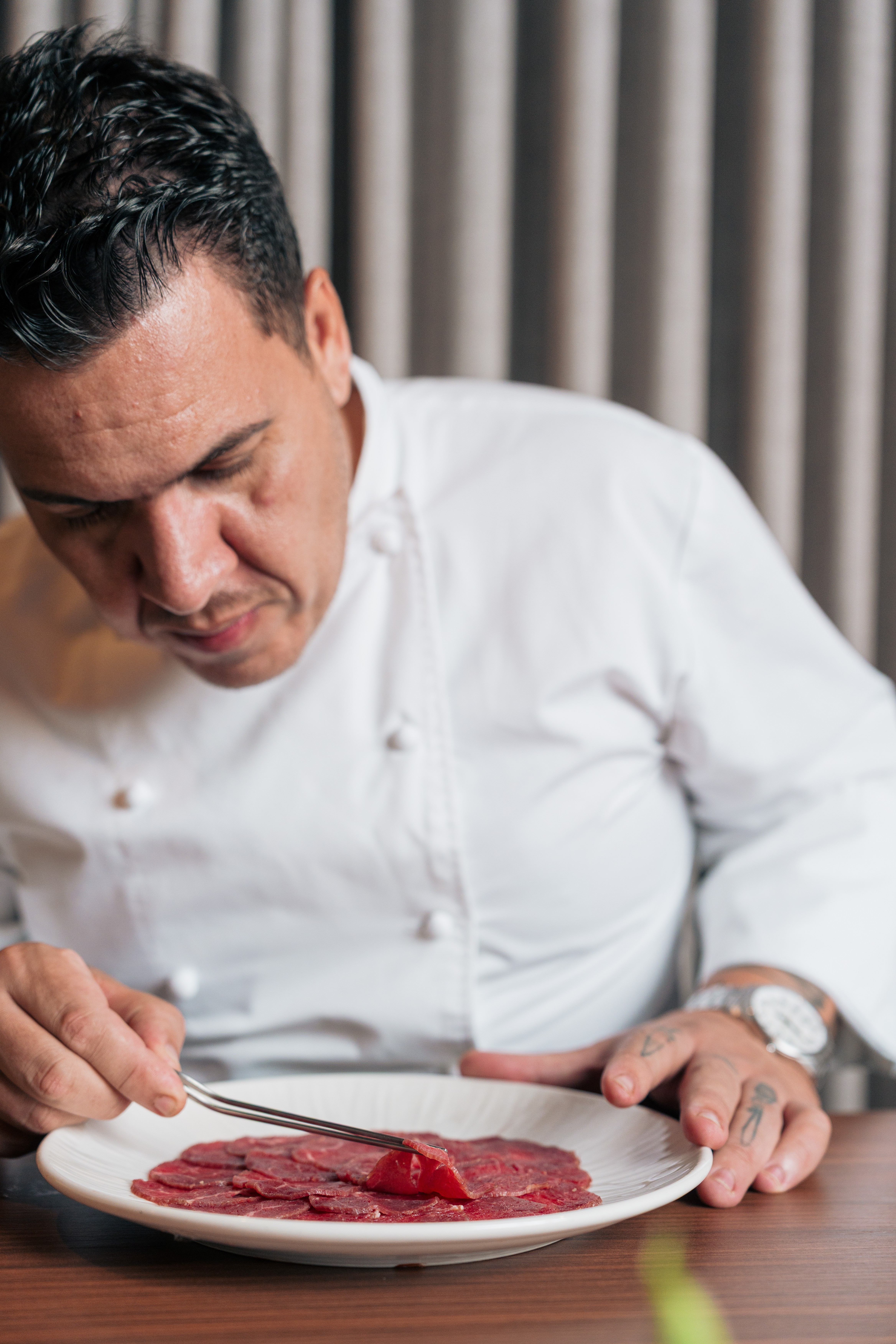 Chef de meia-idade, vestindo d&oacute;lm&atilde; branco, usando pin&ccedil;a para arrumar fatias finas de carne vermelha em um prato branco, sobre mesa de madeira