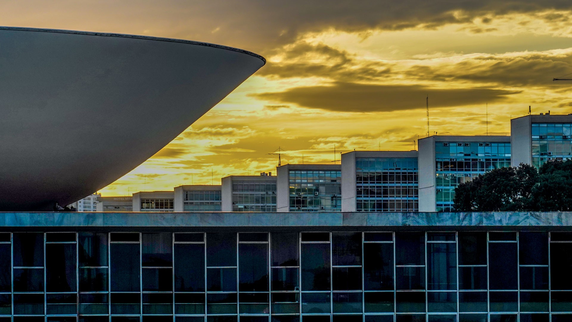 Cúpula côncava do Congresso Nacional em primeiro plano, com edifícios ministeriais ao fundo sob um céu alaranjado e nublado ao pôr do sol