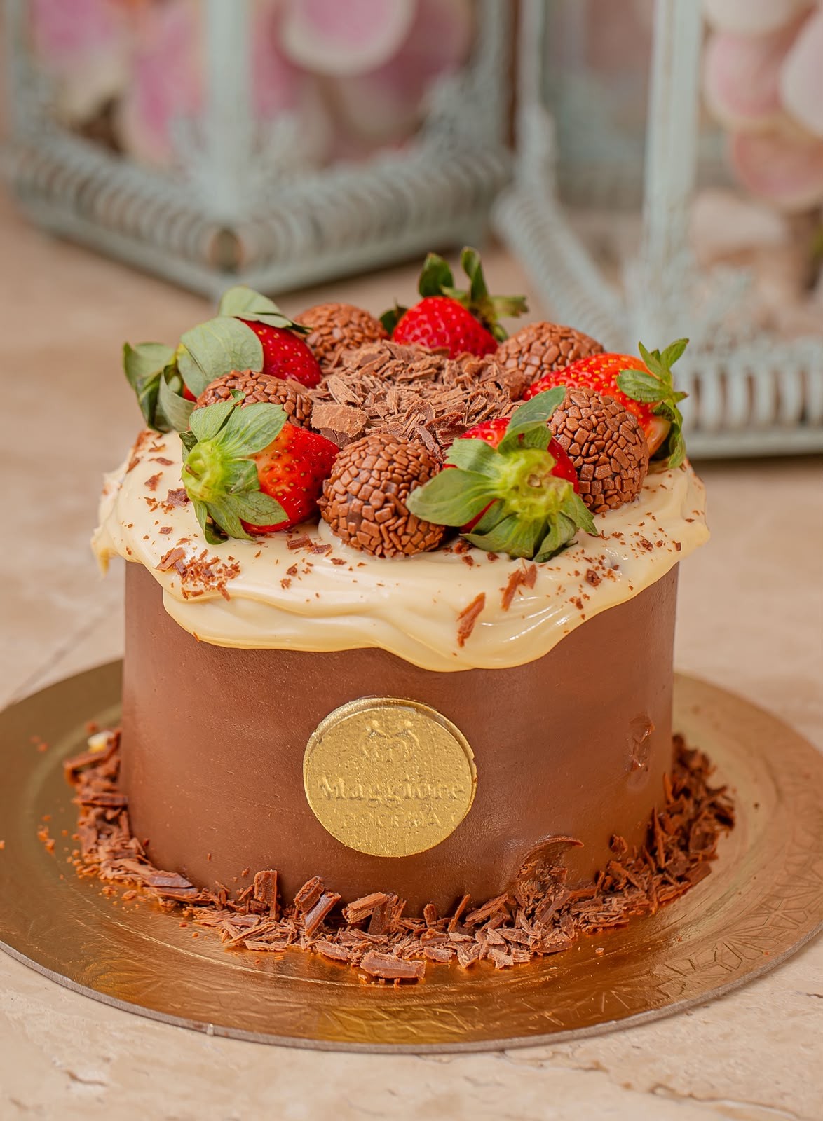 Bolo redondo de chocolate com cobertura de brigadeiro branco escorrendo, decorado com morangos frescos, brigadeiros granulados e raspas de chocolate, sobre um prato dourado. Um selo dourado com o nome Maggiore est&aacute; na lateral do bolo