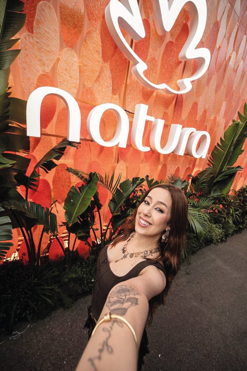 Mulher sorridente de cabelos castanhos e blusa escura, fazendo selfie com braço estendido, exibindo tatuagens e pulseira dourada. Ao fundo, letreiro branco Natura e folha estilizada sobre parede laranja texturizada, com plantas verdes na base