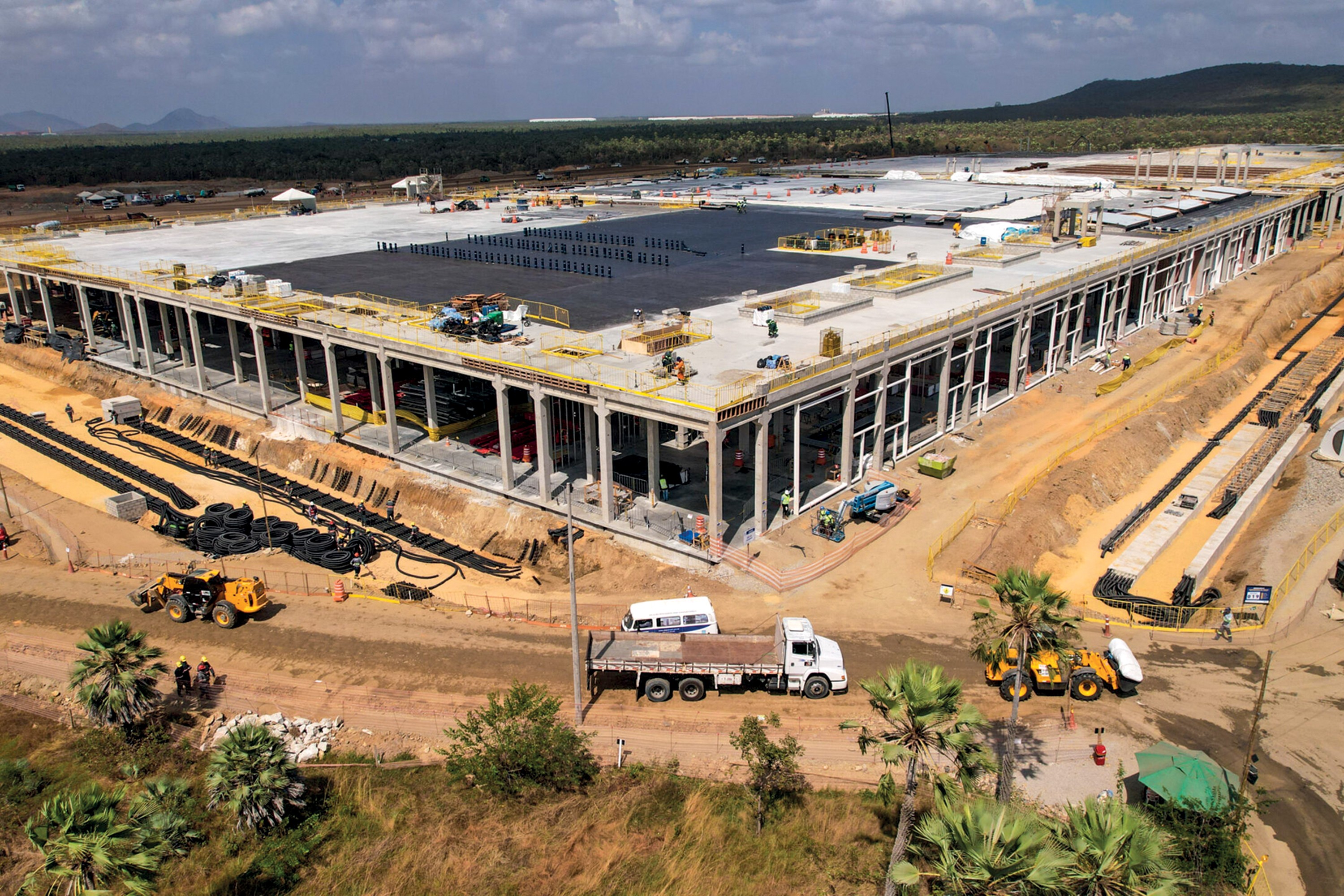 Vista aérea de um grande canteiro de obras com um edifício retangular em construção, com estrutura de concreto aparente e telhado parcialmente coberto. Ao redor, há terraplanagem, máquinas pesadas, pilhas de pneus e materiais, além de caminhões e trabalhadores. Palmeiras e vegetação rasteira pontuam a paisagem.
